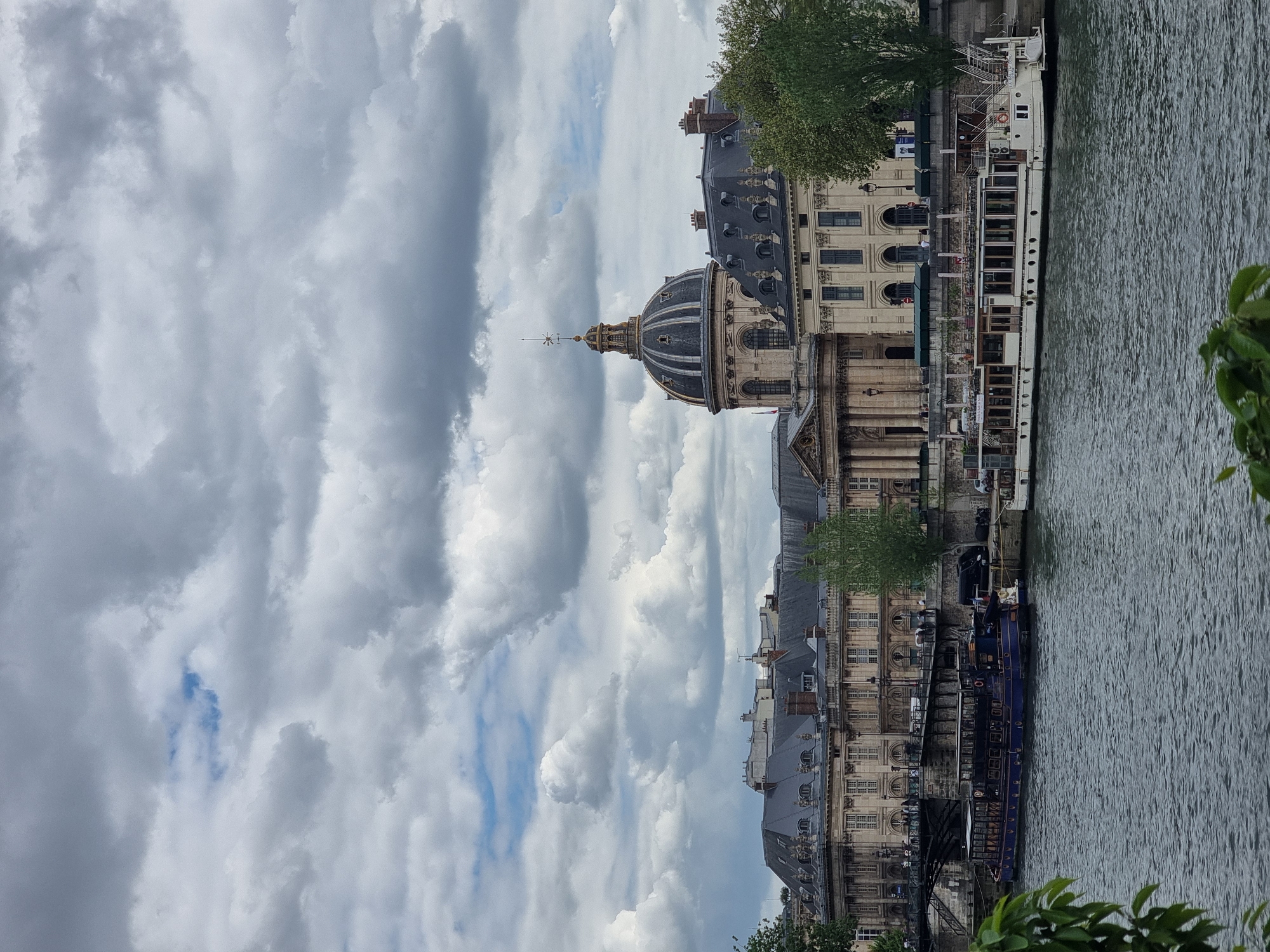 This image captures a picturesque daytime scene along a river in Paris, France, featuring prominent historical architecture under a cloudy sky. The central focus is the elegant dome of the Institut de France, with its distinct classical design and a pointed finial at its apex. This grand building, alongside other stately Parisian structures with classical facades, numerous windows, and grey mansard roofs, lines the riverbank.
The river, likely the Seine, flows in the foreground, its surface rippled under the ambient light. Several boats are docked along the bank. Closest to the viewer, a long, dark blue or purple vessel is moored. Further along, a multi-decked white boat, possibly a tour boat or residential barge, is also docked, showing numerous windows and railings. Indistinct text might be visible on the sides of these boats.
The sky above is largely overcast, filled with a mix of grey and white clouds, although patches of clearer blue sky are visible, suggesting an intermittent break in the cloud cover. Green foliage from trees and bushes frames parts of the scene, particularly on the right side and in scattered spots along the riverbanks, adding natural elements to the urban setting. The overall atmosphere is one of historical grandeur and urban tranquility on an overcast day.