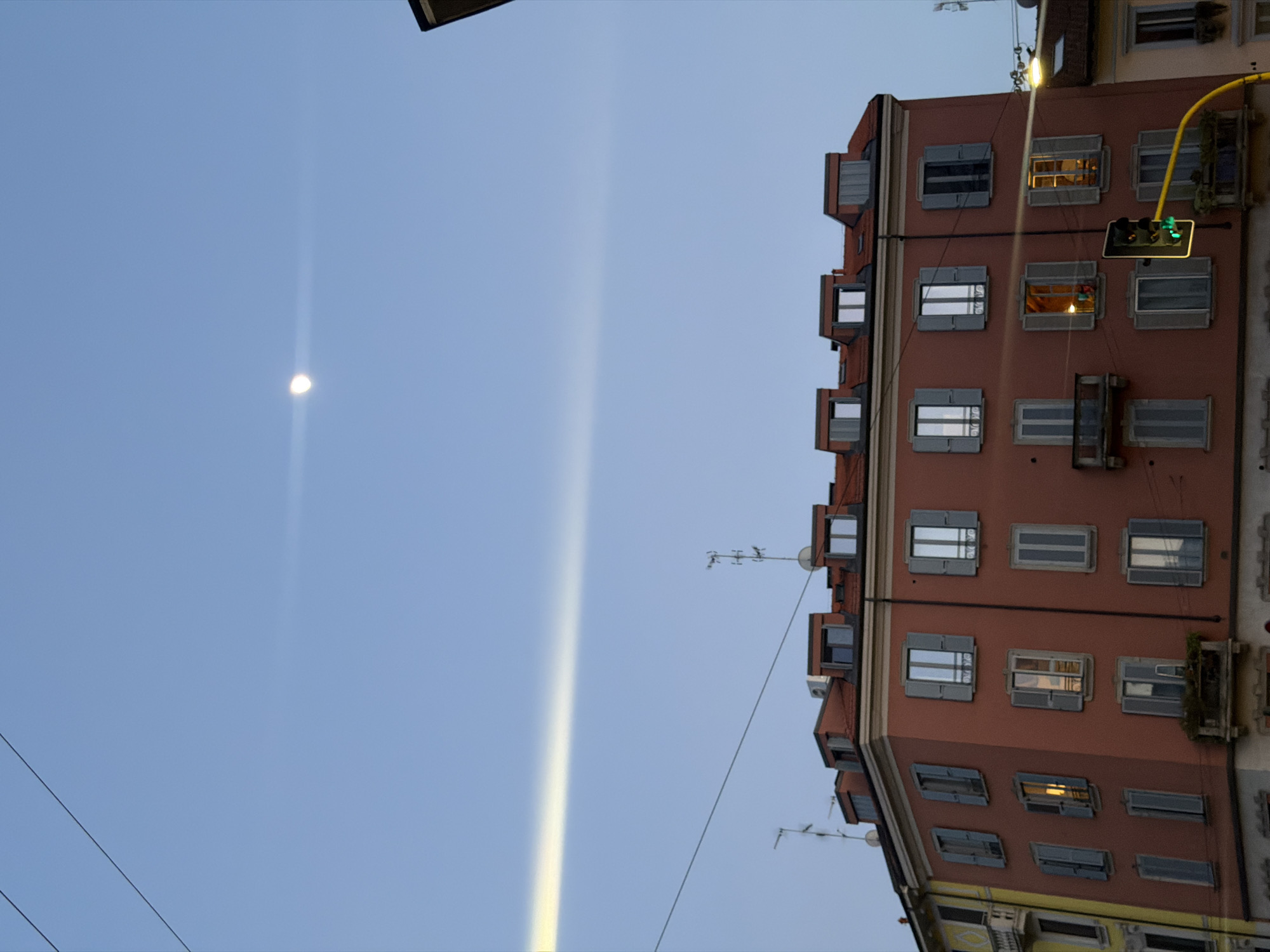 The image shows a view upwards towards the sky in Milan, Italy. A crescent moon is visible in the upper-left portion of the sky. A tall, narrow column of bright light extends vertically from the bottom towards the top. On the right, a multi-story building has windows with gray shutters. A traffic light with green lights is attached to the building. Several antennas are mounted on the roof. Electrical wires traverse the sky.