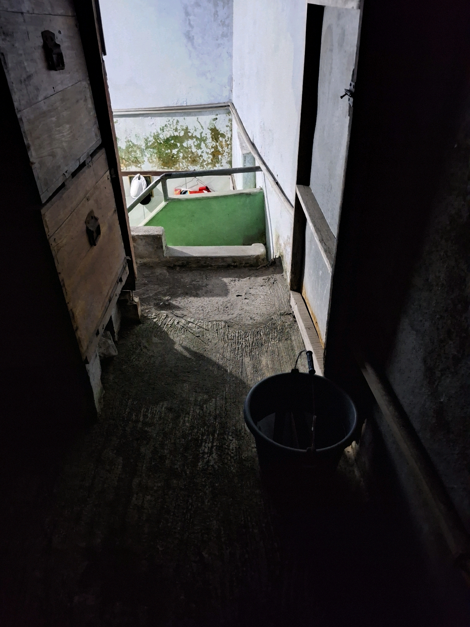 The image shows a dark, gritty alleyway in Lumajang, Indonesia. A dark plastic bucket with a metal handle sits on the wet concrete floor in the foreground, casting a long shadow. To the left, a weathered wooden door is ajar, revealing more of the dimly lit space. Beyond the door, a green, tiled surface suggests a small bathroom or wash area, with a metal railing and a dirty, peeling wall. The overall impression is one of neglect and a lack of maintenance. The lighting suggests it is either nighttime or the area is poorly illuminated.