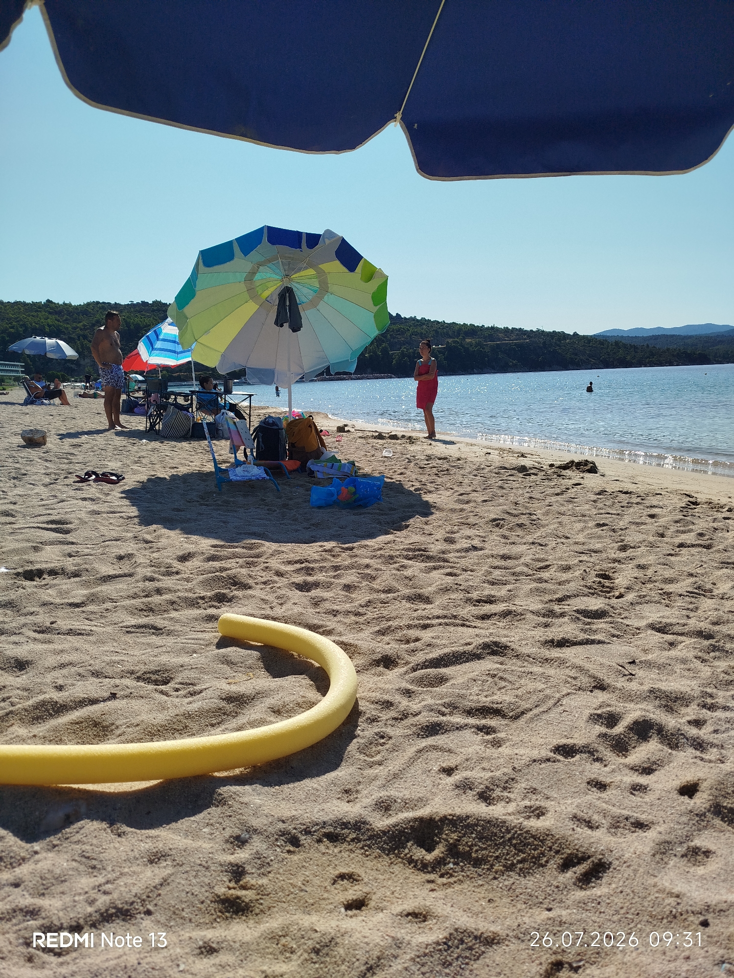 A sunny day at a sandy beach in Sithonia Municipal Unit, Greece. In the foreground, a yellow foam pool noodle lies on the sand. In the mid-ground, several beach umbrellas provide shade. A man stands near a colorful umbrella, while a woman in a red towel stands by the water. Beach chairs and bags are scattered around. The sea is calm and blue, with a distant treelined shore. The sky is clear and bright blue.