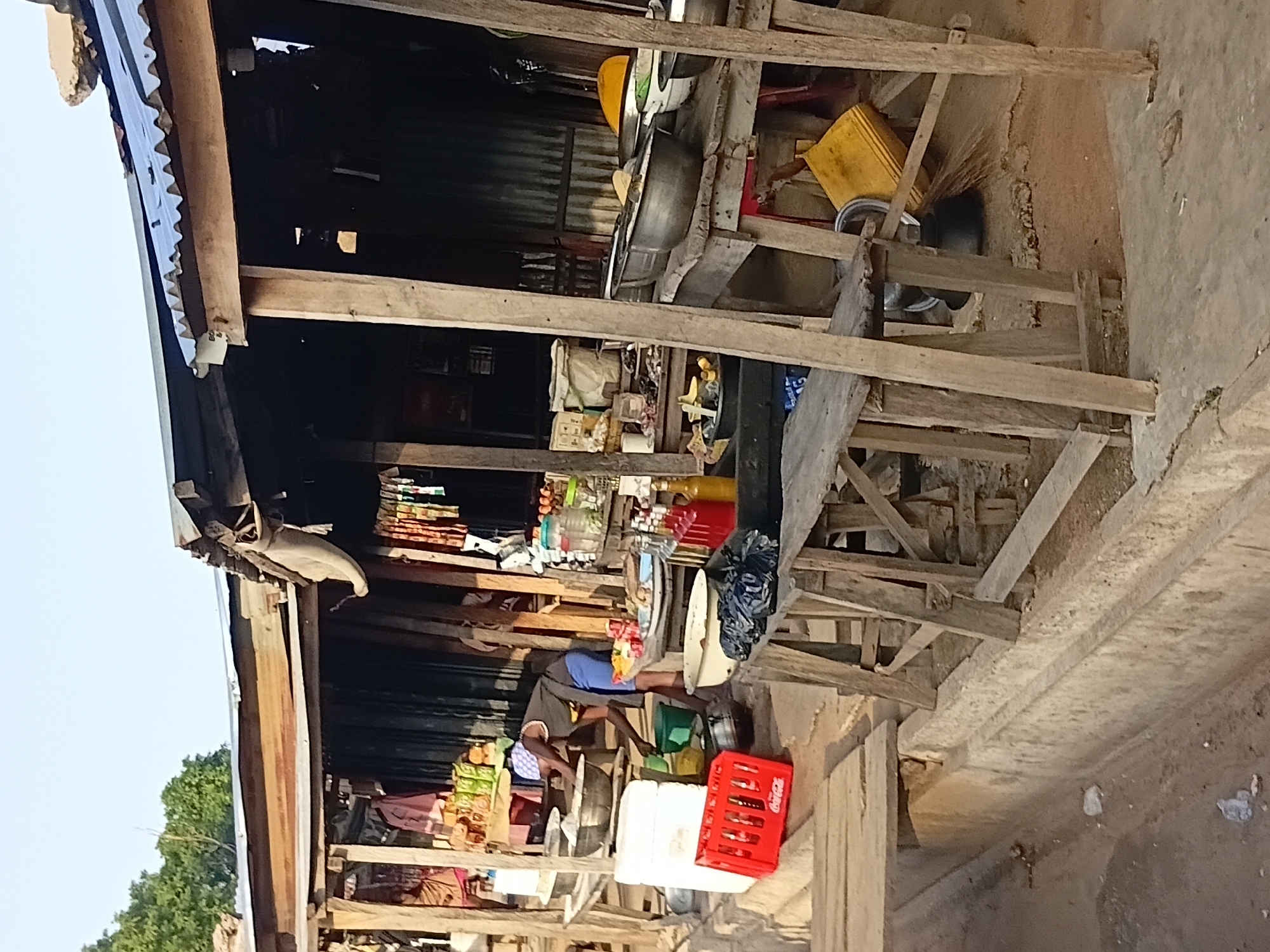 The image depicts an outdoor market stall in Jalingo, Nigeria. The stall is constructed from rough wooden poles and planks, with a corrugated metal roof showing signs of disrepair. Several wooden tables are laden with goods, including what appear to be food items, bottles of liquids, and bags.
In the center of the scene, a person wearing a dark top and blue shorts is bent over, attending to items on a table. They are wearing a checkered head covering. To the left, a red Coca-Cola crate is visible. Large metal bowls and containers are scattered around the stall, along with plastic buckets and what looks like a broom leaning against a wooden support. The ground is a mixture of dirt and concrete. The overall impression is of a busy, somewhat makeshift market environment.