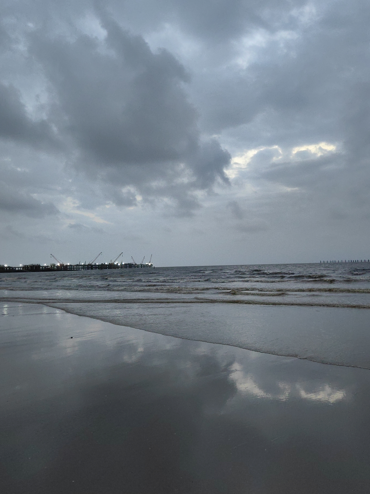 The image depicts a wide shot of a beach in Mumbai, India, under a heavily overcast sky. The foreground features wet sand reflecting the muted light and the sky. Gentle waves lap at the shore, creating a rippling effect on the sand. In the middle ground, the sea stretches towards the horizon, with several rows of waves visible. In the distance, a long pier or jetty extends into the water. The pier is lined with numerous lights, and several tall cranes are positioned along its length, some of which are illuminated. Further out at sea, a series of pilings or posts mark the edge of the water. The overall atmosphere is somber and industrial, with the cloudy sky and the utilitarian structures in the distance dominating the scene. There are no people visible in the image.