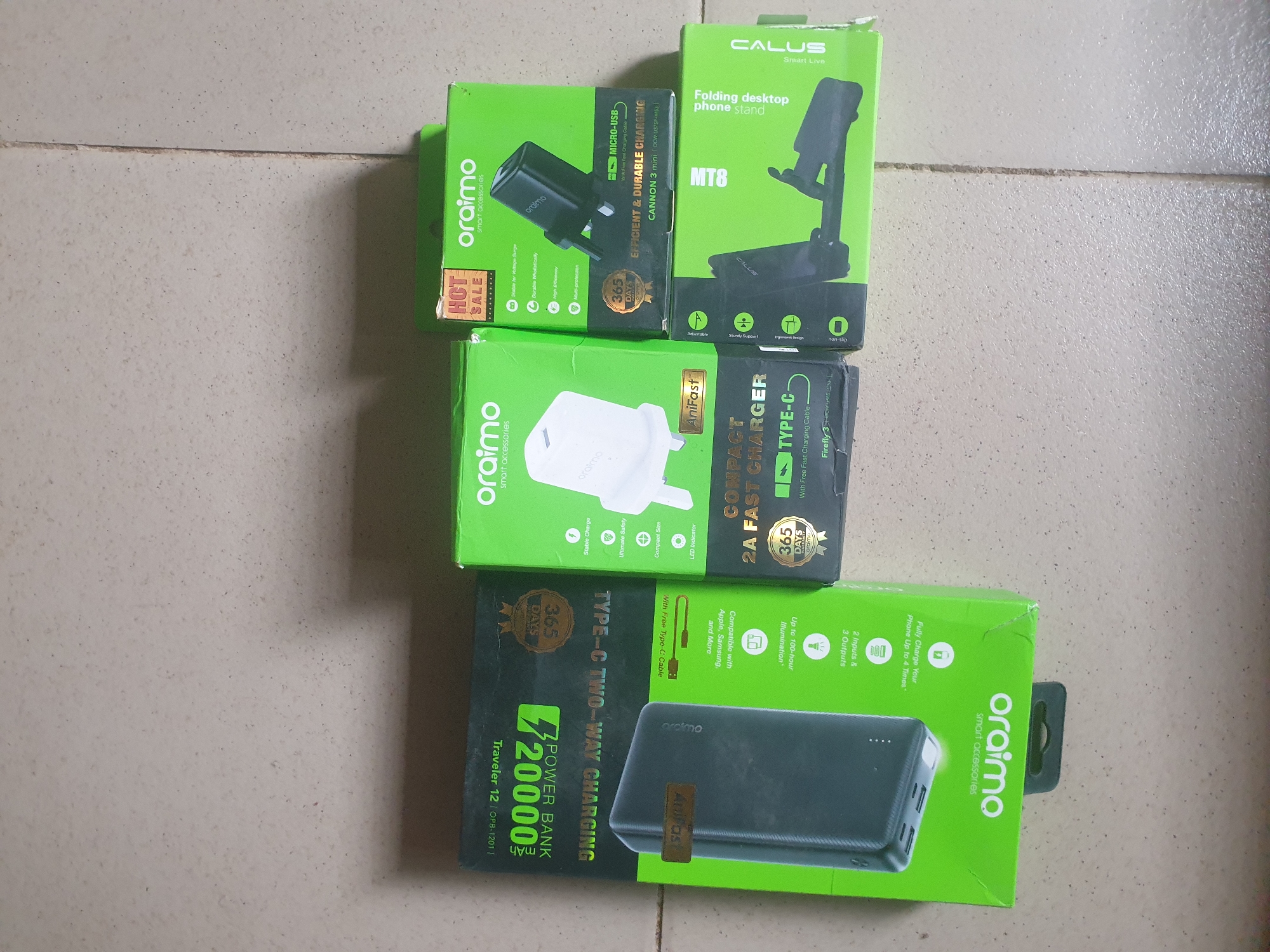 In Anilelerin, Nigeria, a collection of electronic accessories is displayed on a light-colored, textured surface, possibly a wall or floor. The items are arranged on top of their respective product packaging.
In the foreground, to the left, is a dark gray power bank, partially visible within its bright green and black packaging. The packaging prominently features the "oraimo" logo and text indicating "20000mAh POWER BANK" and "TYPE-C TWO-WAY CHARGING."
To the right of the power bank, several Orlaimo brand smart accessories are presented in green boxes. One box, with a "HOT SALE" sticker, shows a black power adapter with the "oraimo" logo. Text on this box indicates "EFFICIENT & DURABLE CHARGING" and "MICRO-USB." Another Orlaimo box to its left contains a white power adapter, also branded "oraimo" and featuring "AniFast" technology.
Further to the right, another Orlaimo green box displays a black charging plug and text related to "EFFICIENT & DURABLE CHARGING." Below these, a green box for a "CALUS" brand folding desktop phone stand is visible, with the product itself also shown.
The overall scene depicts a retail display or collection of new electronic products, suggesting a commercial setting within Anilelerin.