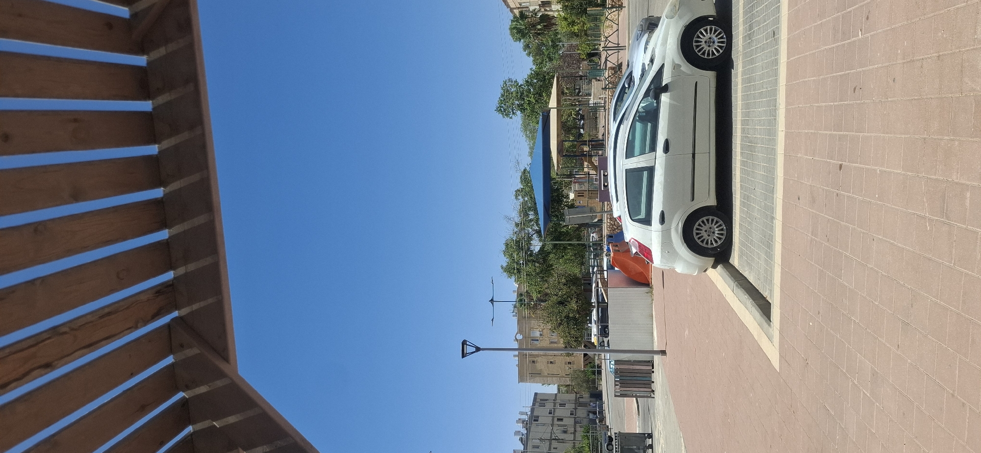 The image depicts a street scene in Yerocham, Israel, likely during daylight hours given the bright blue sky and shadows. In the foreground, a white Fiat Punto is parked in a paved lot. The car is facing towards the right of the frame. Behind the white car, another vehicle, appearing to be a silver sedan, is partially visible.
The street itself is paved with reddish-brown bricks, leading to a darker grey tiled parking area where the cars are parked. A curb separates the sidewalk/parking area from the street. To the left of the parked cars, a modern lamppost with a square head stands prominently. Further in the background, there are buildings, trees, and what appears to be a playground area with a blue canopy and a beige structure. A slide is visible on the left side of the playground. Several other vehicles and structures are scattered throughout the background, suggesting an urban or suburban environment.
The overhead view reveals a wooden pergola-like structure partially framing the top of the image, casting shadows on the blue sky. The overall impression is one of an outdoor public space in a warm climate.