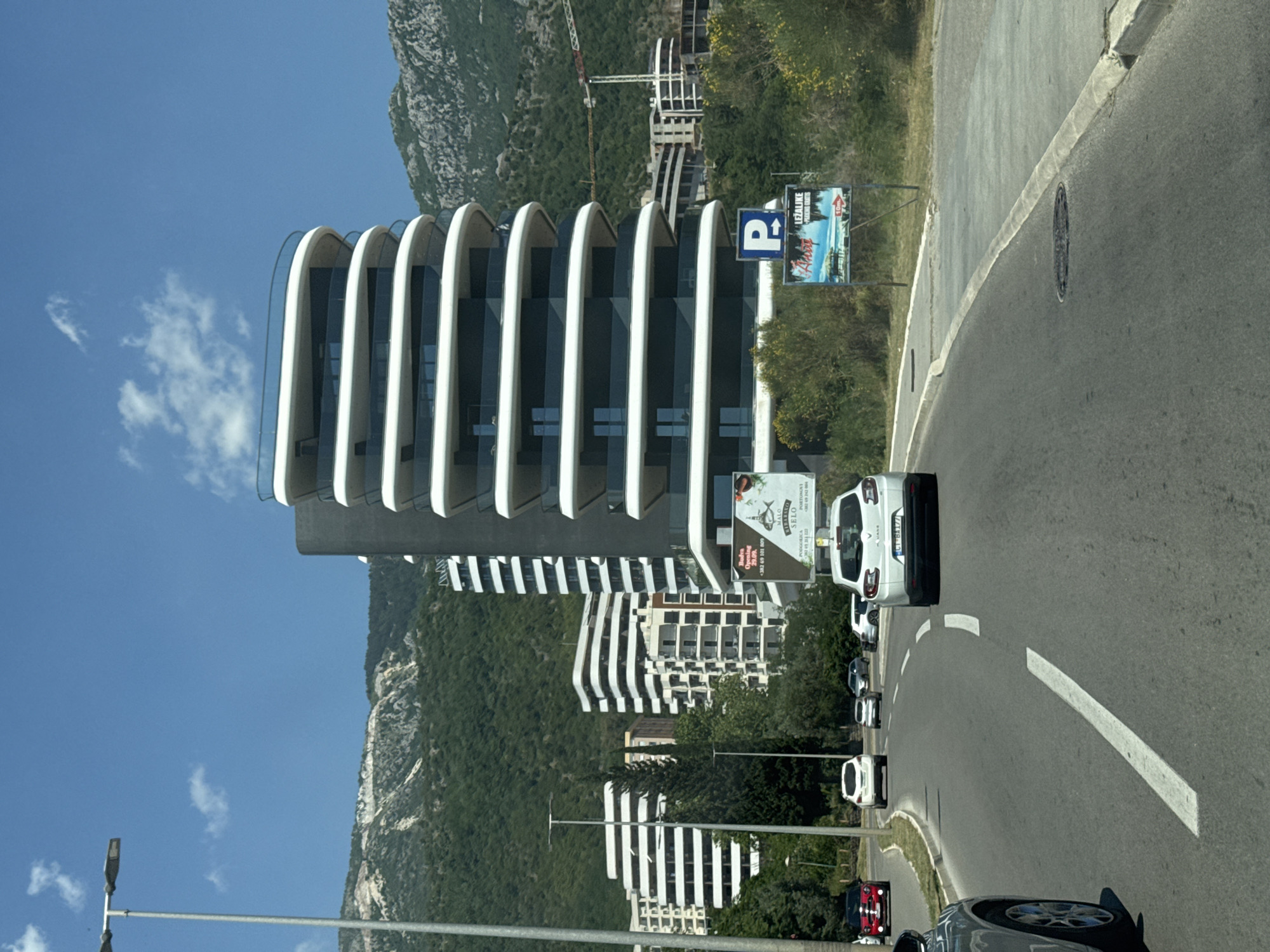 The image captures a modern architectural scene near the city of Boreti, Montenegro. A multistory building with a unique design featuring horizontal white curved elements stands prominently, backed by a mountain range. A road curves in front of the building with several parked cars. Signage is visible, including a parking sign with a "P" on it. Another sign shows opening details for "MALO RIBARSKO SELO". The sky is mostly clear blue.