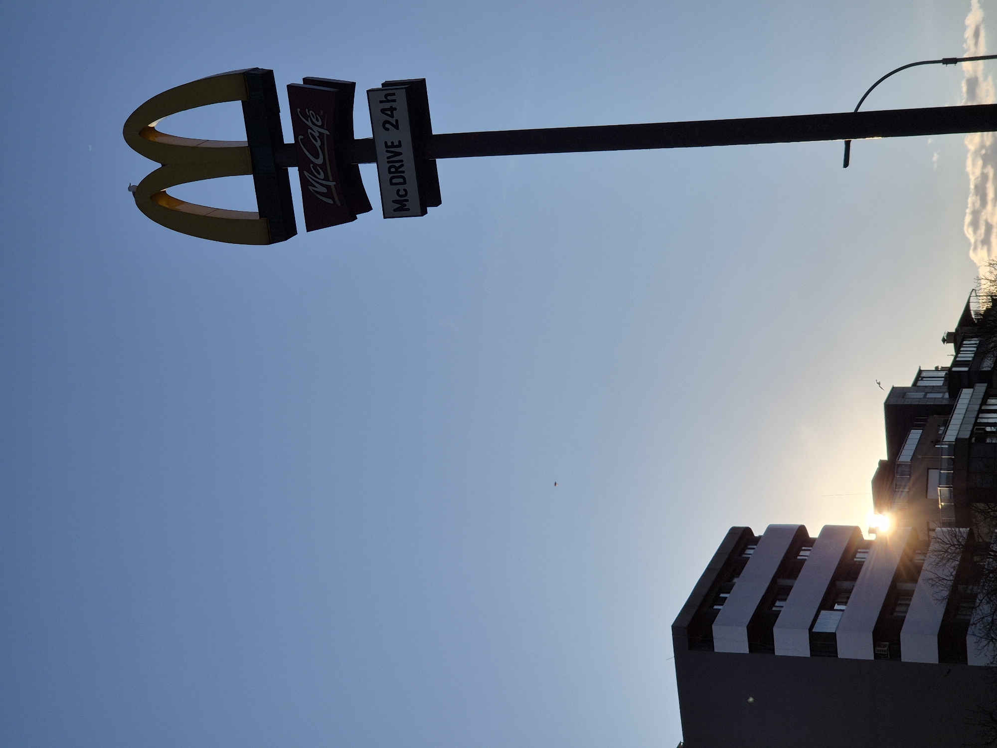 The image is taken from a low angle, looking up at a McDonald's sign against a clear blue sky. The sign includes the golden arches logo, a "McCafé" sign, and a "McDrive 24h" sign. The sign is mounted on a tall, black pole. In the lower right corner, a modern building with a distinct tiered design is visible, with the sun shining behind it. The location is Varna, Bulgaria, based on the architectural style of the building and the presence of the McDonald's sign within the urban environment. The environment appears to be urban, based on the building and signage. There are no people visible.