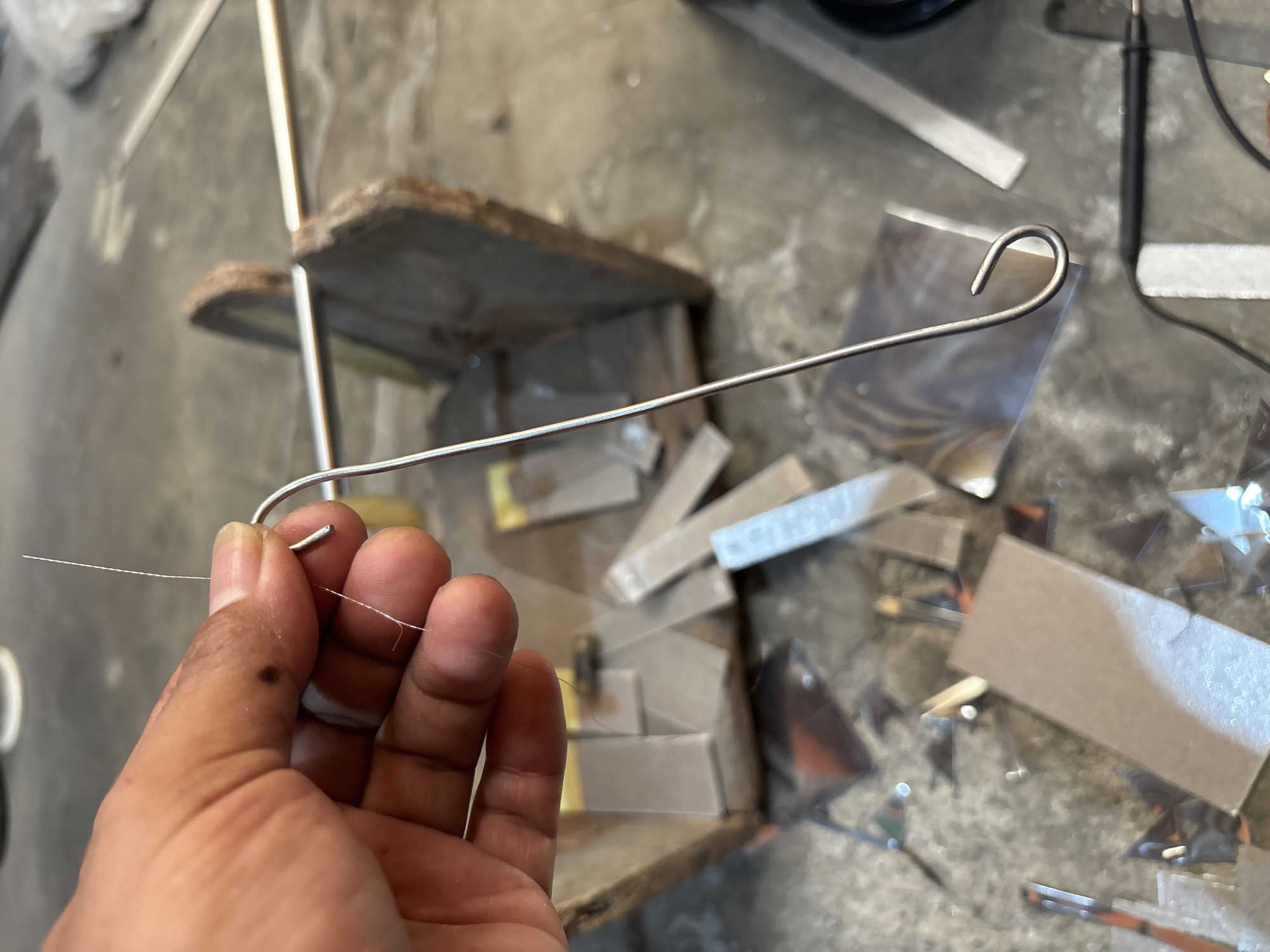 A close-up shot captures a person's hand holding a thin, metallic hook-like object. The hand, with visible skin texture and fingernails, gently grips a fine, translucent filament extending from the hook. The background suggests a workshop or a cluttered workspace, with various materials scattered on the floor, including what appear to be pieces of glass or reflective material, and wooden objects. The overall impression is one of manual work or crafting, possibly involving delicate assembly or repair. The location of Telagamurni, Indonesia, is not discernible from the visual information provided in the image.