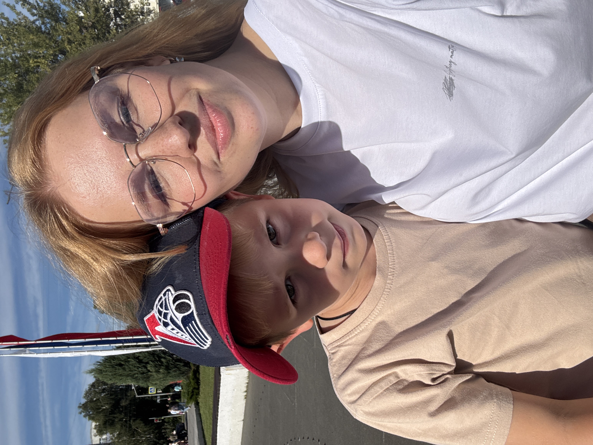 In Rusanovo, Russia, on a bright, sunny day, a young boy and a woman are taking a selfie outdoors. The boy, wearing a beige t-shirt and a blue and red baseball cap with a logo, is positioned to the left of the frame, looking towards the camera with a neutral expression. The woman, with blonde hair and wearing a white t-shirt and glasses, is on the right side of the frame, smiling slightly at the camera. They appear to be standing on an asphalt surface, with a white line on the ground in the foreground. In the background, there are trees, a building, and what appears to be a flag pole with a flag. The overall setting suggests an outdoor public space.