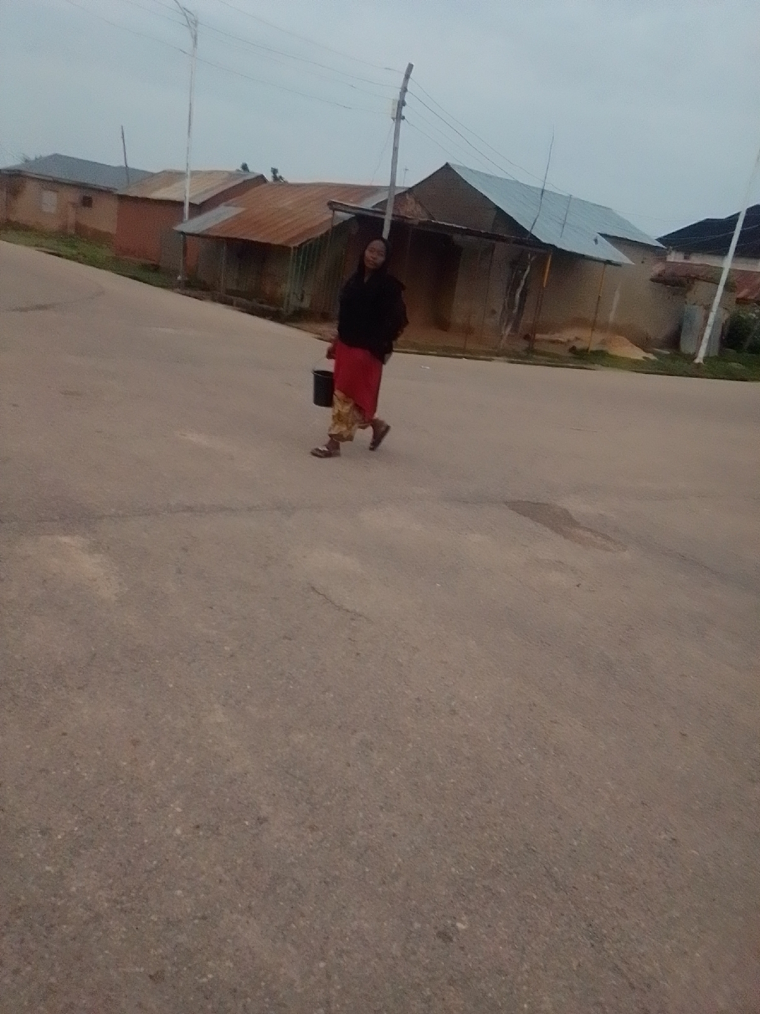 A person walks on a paved road in Gombe, Nigeria, carrying a black bucket. The person is wearing a dark top and a patterned skirt in shades of red and yellow. Buildings with corrugated iron roofs and concrete walls are visible in the background, along with utility poles and power lines. The sky is overcast.