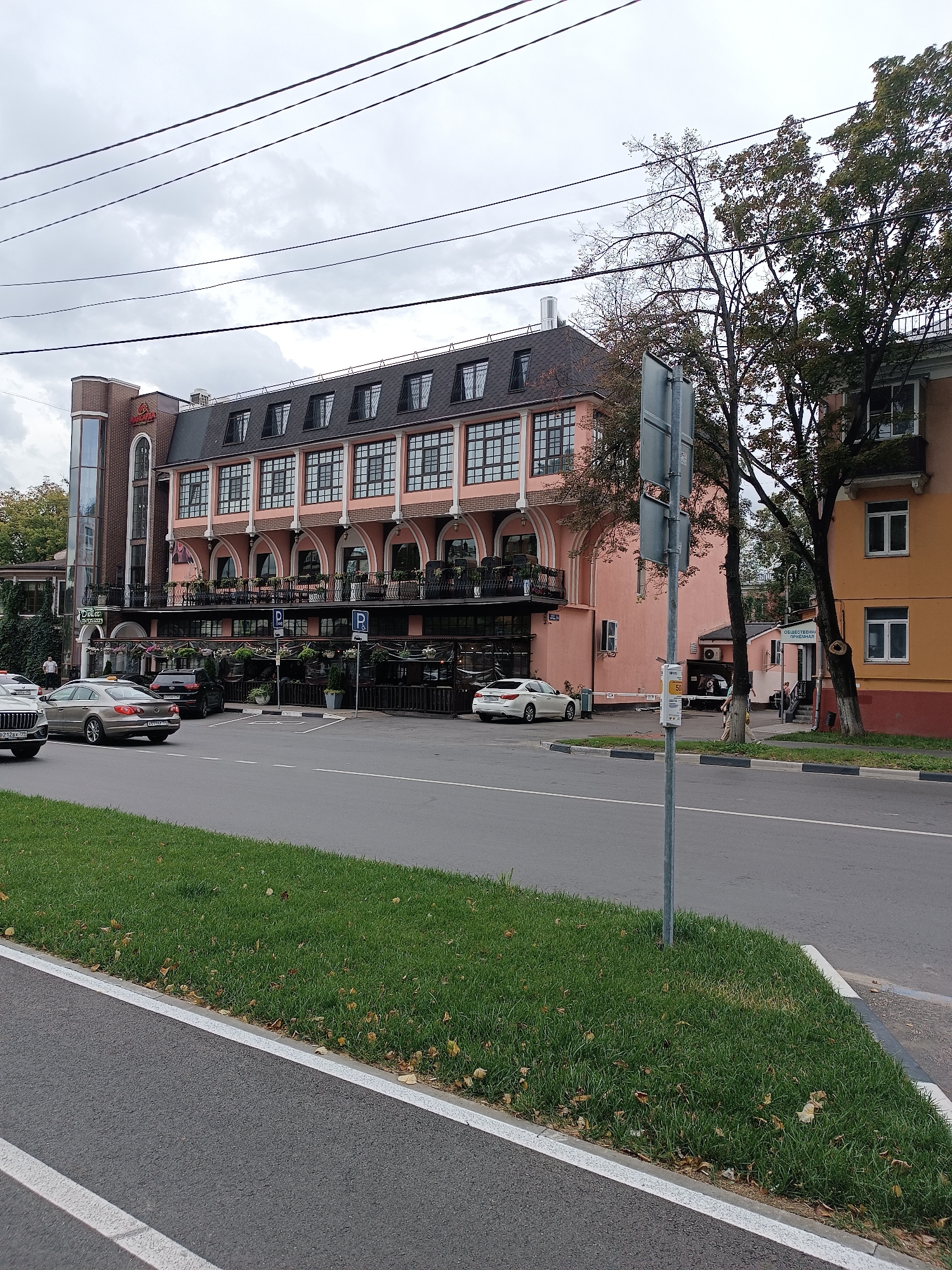 The image depicts a street scene in Lyubertsy, Russia, with a prominent pink building on the right side of the frame. The building features multiple floors with arched windows and balconies, and a dark-colored mansard roof. Several trees with green and yellowing leaves are visible in front of and beside the building, indicating an autumn season. A street with traffic runs in the foreground, with a white car in the left lane and a gold sedan in the right lane. A black SUV is parked near the building, along with other vehicles. A traffic signpost stands on the right side of the road. The sky is overcast and grey. The overall impression is that of a commercial or residential area on a cloudy day.