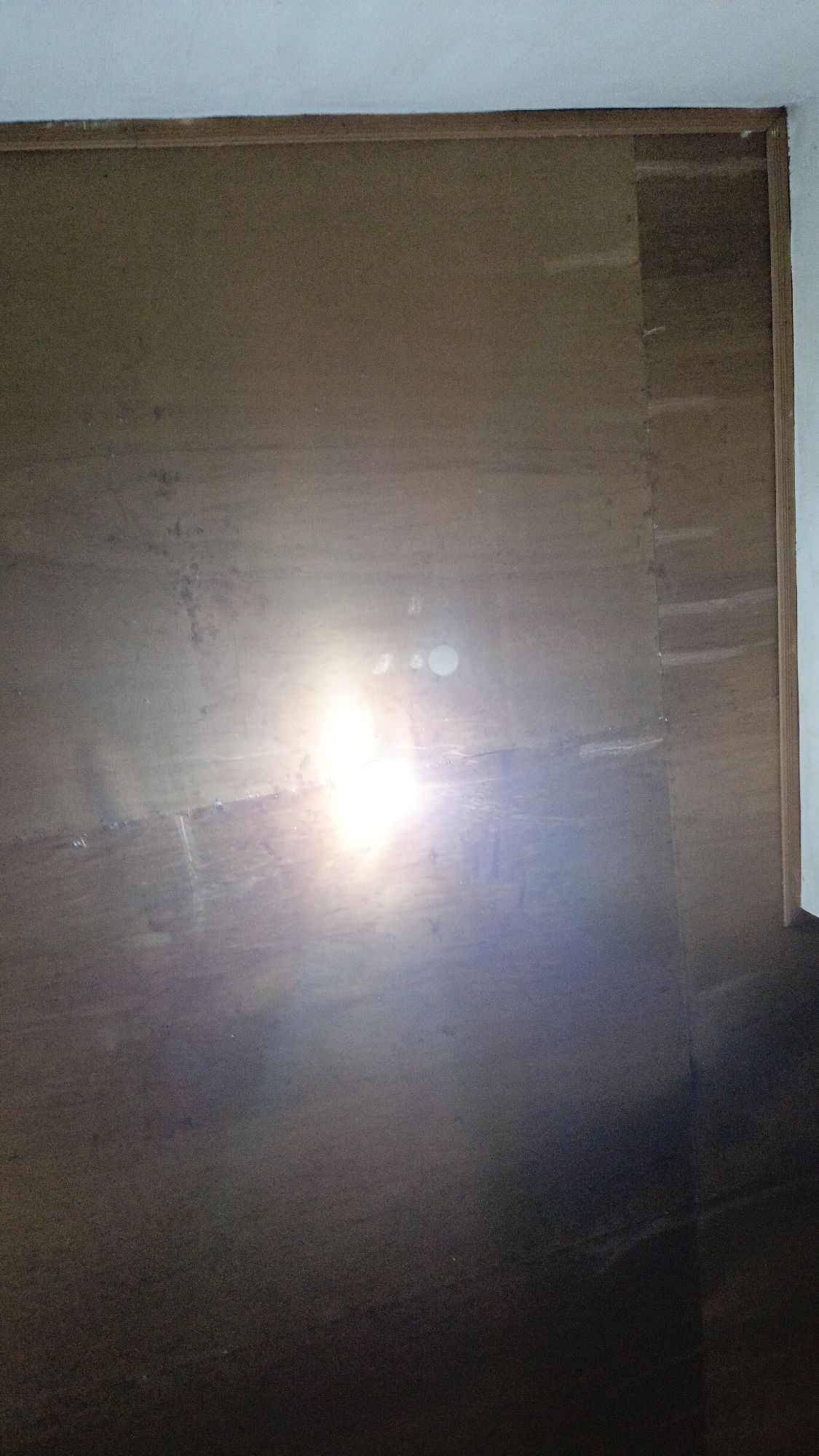 The image displays a close-up, low-angle shot of a dark, textured surface that appears to be made of wood or a similar material. The surface is divided into several sections by vertical lines, suggesting it might be a door, a cabinet, or a panel. A bright light source, possibly a flashlight or camera flash, is prominently reflecting off the surface in the center of the image, causing a strong glare and lens flare. The light creates a halo effect and illuminates some of the texture and imperfections on the surface, such as scuff marks and subtle variations in color. The top edge of the image shows a sliver of a white wall and a thin wooden frame. The lighting suggests an indoor environment, likely dimly lit apart from the direct light source. There are no visible people or specific landmarks to suggest a precise location within Kota Pagatan, Indonesia. The overall impression is of a utilitarian, possibly weathered, surface.
