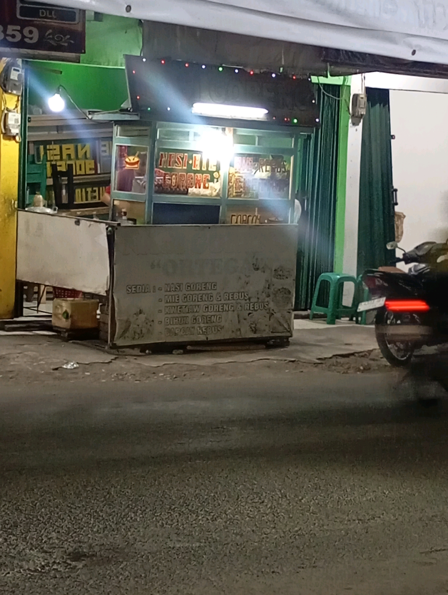 The image shows a street-level view of a food stall in Babelan, Indonesia. The stall appears to be open for business at night, with bright lighting illuminating the area. The stall is covered by a green awning and has a display window with food items visible. There are signs with text written in Indonesian, some of which are legible, indicating that it serves dishes like "Nasi Goreng" (fried rice) and "Mie Goreng" (fried noodles). In front of the stall, there are two green plastic stools and a motorcycle parked to the right. The road in front of the stall is paved and appears to be a public street. The overall scene depicts a typical roadside food vendor operating in an urban environment.
