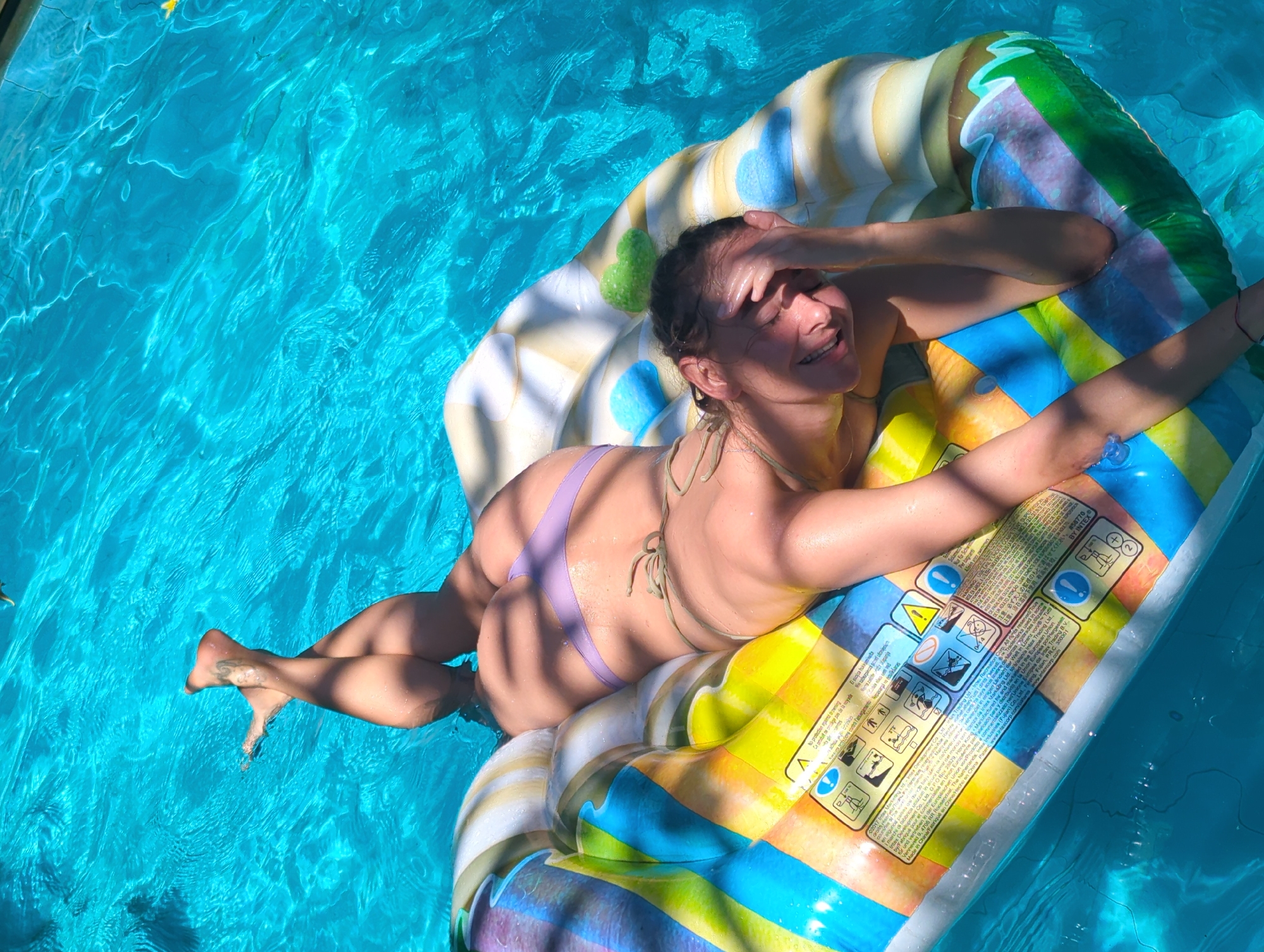 A woman is lying on an inflatable raft in a swimming pool filled with turquoise water. The raft is brightly colored with stripes and printed warnings. She is wearing a light purple bikini and has her right hand over her eyes. Her left arm is extended outward. The pool water reflects sunlight.