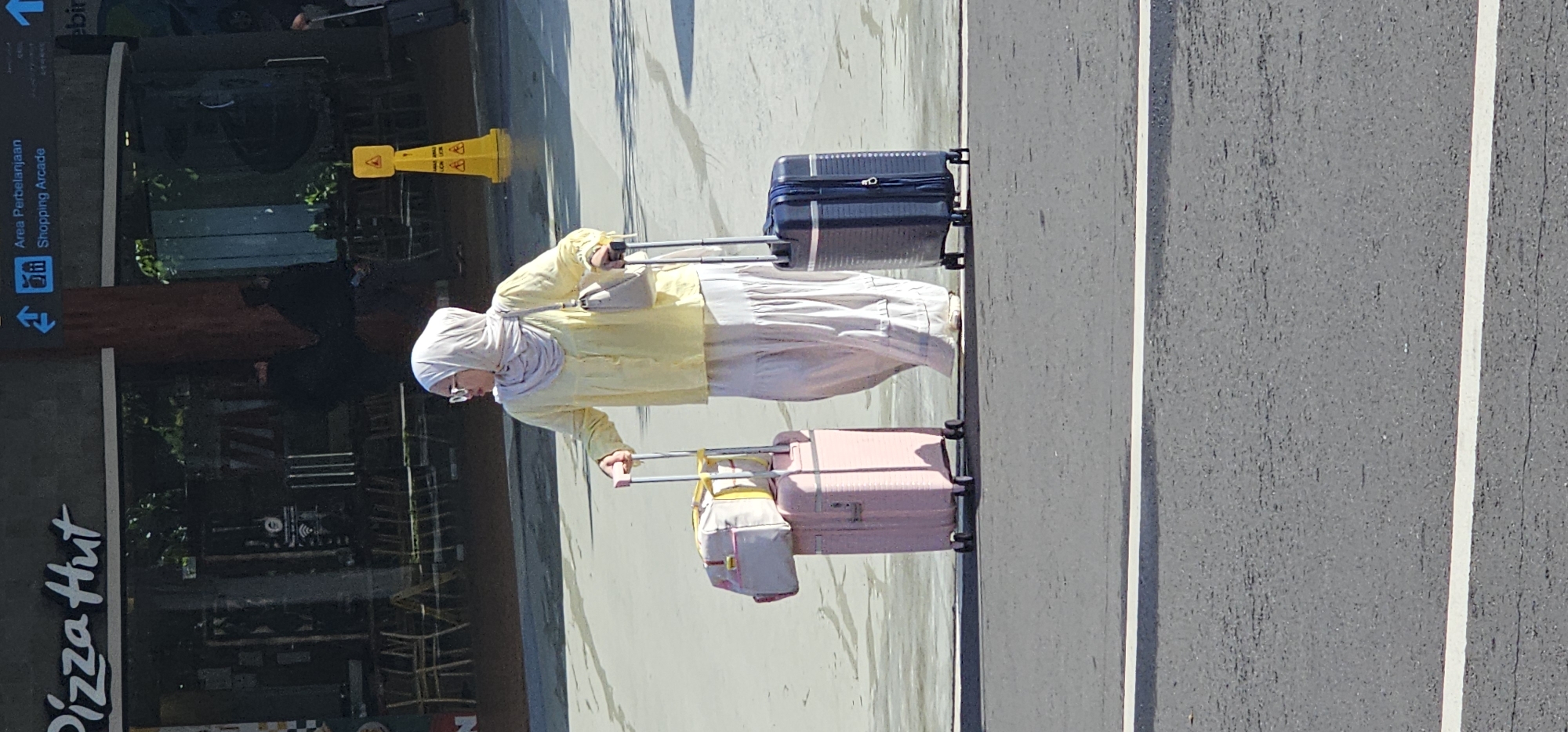 A woman is standing on the road between two white lines, pulling two suitcases. The woman is wearing a hijab and glasses, with a yellow shirt and a cream-colored pants. One suitcase is dark and one is pink. The sign for Pizza Hut can be seen in the background. A sign for a shopping arcade can also be seen. This picture appears to be taken in Kuta, Indonesia.