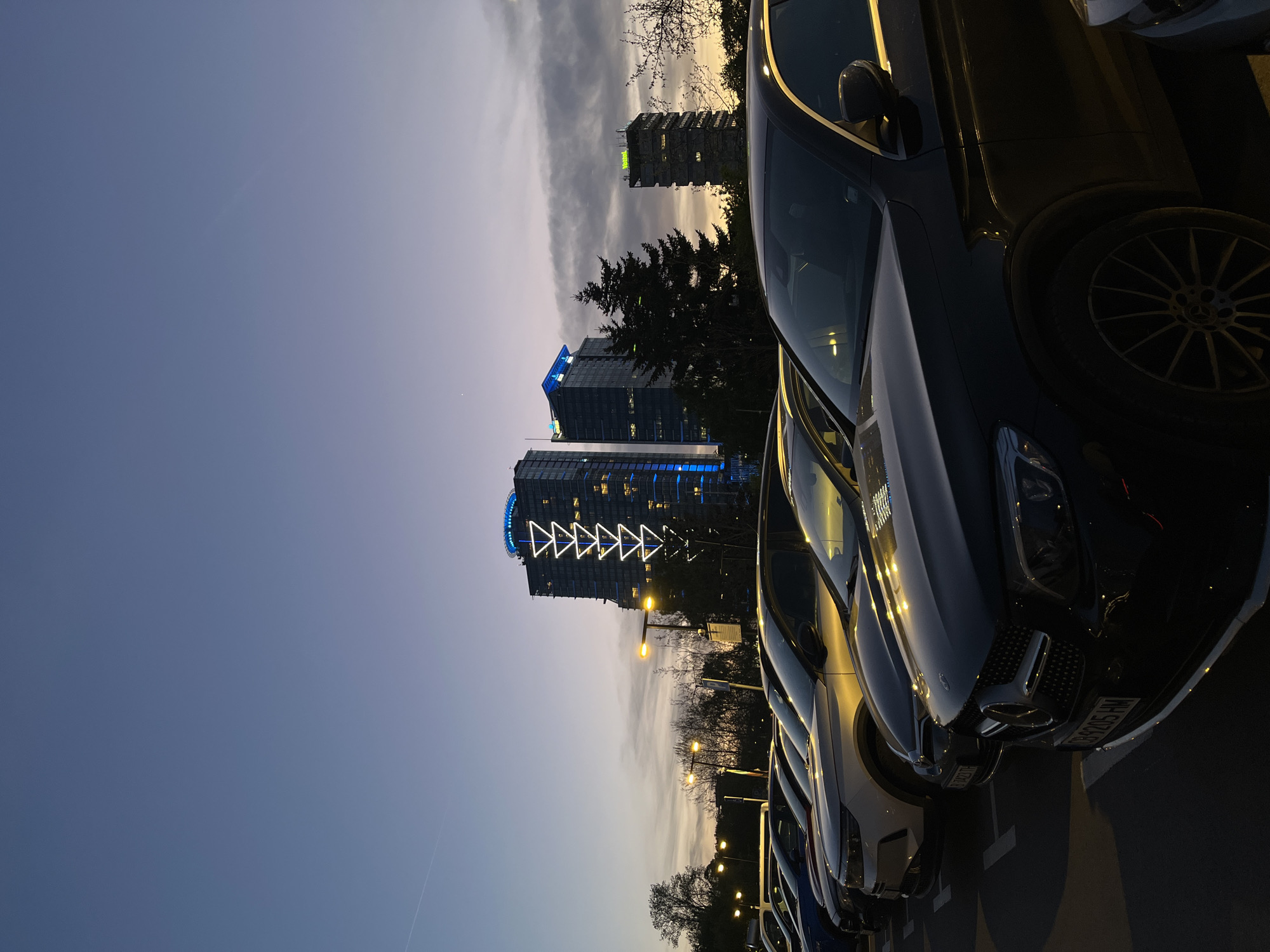 The image, correctly oriented, captures an urban street or parking lot scene in Sofia, Bulgaria, during dusk or early evening. In the foreground, a line of parked cars is visible, with a dark-colored Mercedes-Benz occupying the right side of the frame. Its front grille, headlights, and a multi-spoke wheel are discernible, along with reflections from the ambient light on its body. The license plate "CB 9205 HM" is visible on the front of this car.
Behind the vehicles, several yellow-orange streetlights illuminate a section with trees. The background is dominated by modern high-rise buildings. One prominent building on the left features distinctive blue lighting and an elaborate illuminated white triangular pattern on its exterior. Another building adjacent to it also displays blue illumination. Further to the right, a more distant building appears to have green lighting at its peak.
The sky above is in twilight, transitioning from a lighter tone on the right to a deeper blue towards the left, indicating the fading daylight. Scattered clouds are present, and faint, linear contrails, likely from an aircraft, are visible in the upper sky. The overall scene conveys a calm, modern urban environment as night descends. No people or specific activities are depicted; the focus is on the city's infrastructure and architecture.