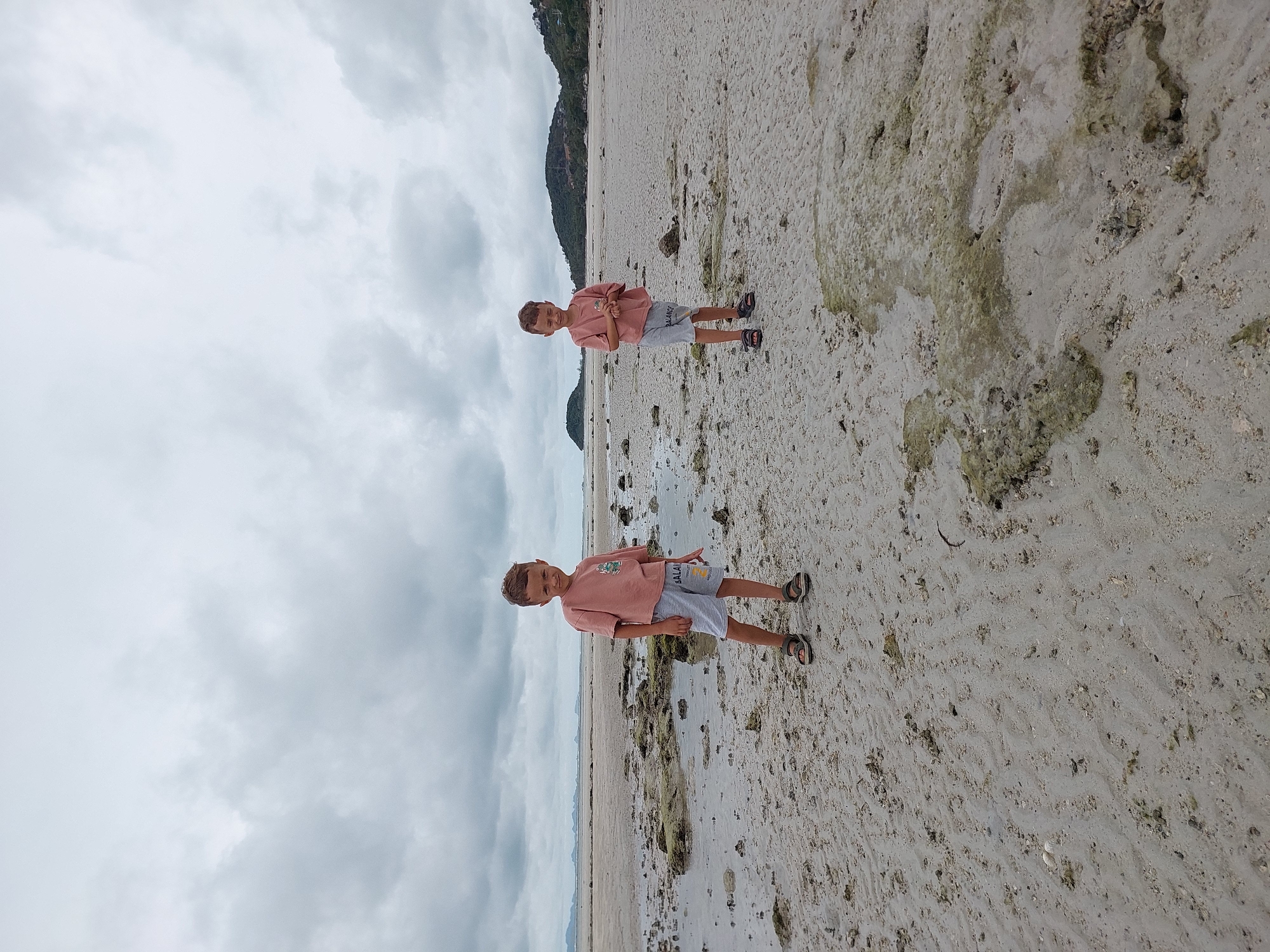 The image depicts two young boys standing on a wet, sandy beach with cloudy skies overhead. The boy in the foreground is wearing a pink t-shirt and grey shorts, with sandals on his feet. He is looking towards the camera with a slight smile. His body is positioned at an angle, with his hands clasped in front of him. Behind him, another boy, dressed identically, is partially visible, also standing on the beach. The sand is wet and textured, with some small rocks and patches of green algae visible. The background shows a faint outline of hills or a mountainous landscape under a dramatic, overcast sky. The overall lighting suggests it is either early morning or late afternoon. The setting appears to be a tropical beach, with the mention of Ko Samui District, Thailand providing specific location context. There is some visible text on the boys' shorts, though it is not clearly legible.