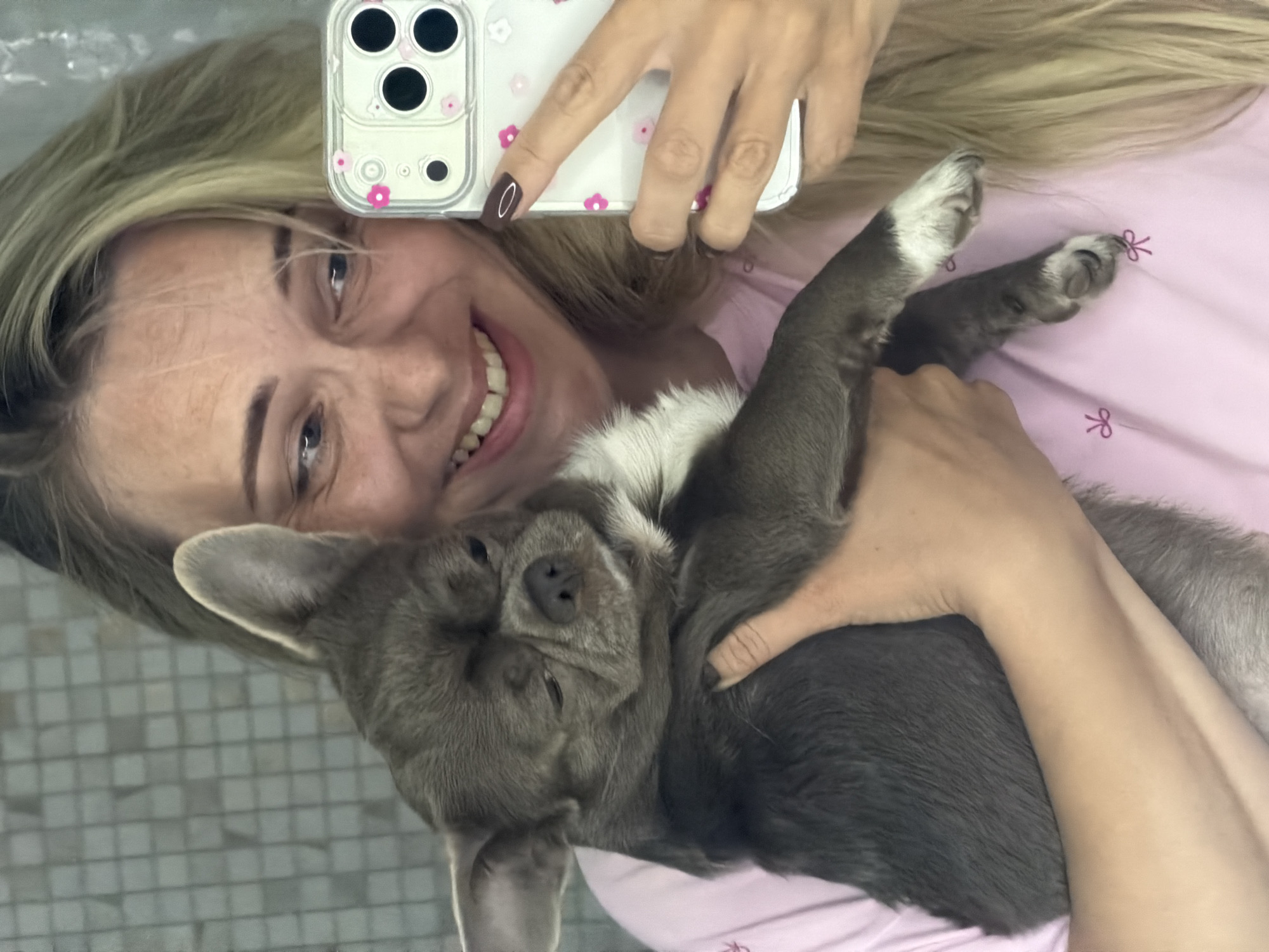 A blonde woman with fair skin, wearing a pink t-shirt with small embroidered bows, is taking a selfie with her grey chihuahua. The dog is held in her left arm, its face tilted upwards with closed eyes. The woman is smiling, looking at her phone held in her right hand. Her fingernails are painted a dark brown. The background appears to be a tiled wall, possibly in a bathroom. The image was taken in Votkinsk, Russia.