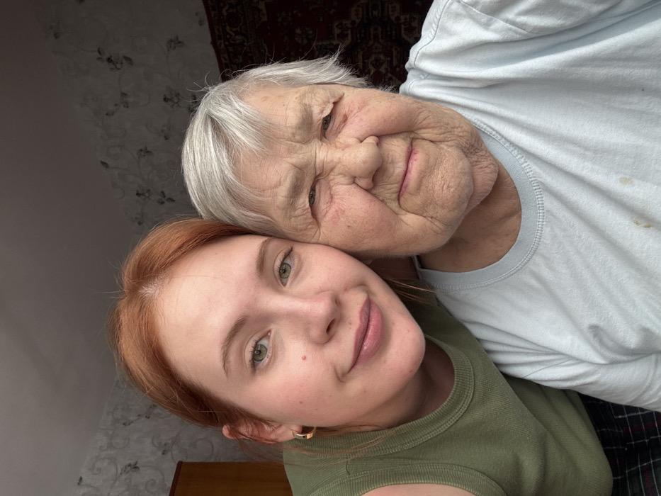 The image displays a close-up, indoor portrait of two females of differing generations, photographed within a residential setting in the city of Chaikovsky, Russia. The individual on the left is a younger adult female with reddish-brown hair, fair skin, and green eyes, wearing an olive green ribbed tank top and a gold-colored hoop earring on her visible left ear. She exhibits a subtle smile. Adjacent to her on the right, positioned cheek-to-cheek, is an elderly female with white-grey hair, significantly wrinkled fair skin, and a neutral facial expression. She is dressed in a light blue crew-neck t-shirt, which bears faint yellowish-brown stains on its lower visible area. The background elements include light-colored wallpaper adorned with a subtle floral pattern, a partial view of a wooden wardrobe or cabinet on the far left, and a dark, patterned textile (possibly a rug or tapestry) hanging on the wall to the far right.