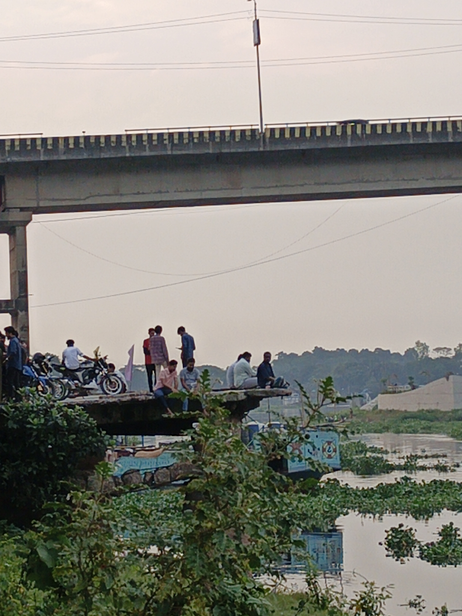 The image depicts a group of people gathered on what appears to be a concrete structure near a body of water, possibly a river, in an unknown city. The structure, which is situated close to the water, supports several individuals, some of whom are standing, while others are seated. Two motorcycles are parked on the structure. The water is covered with vegetation, possibly water hyacinth, and there is a barge in the water. Above the group and the structure is a large bridge that spans the water. A light pole is seen on the bridge. The background includes trees and buildings. The scene is captured in daylight.
