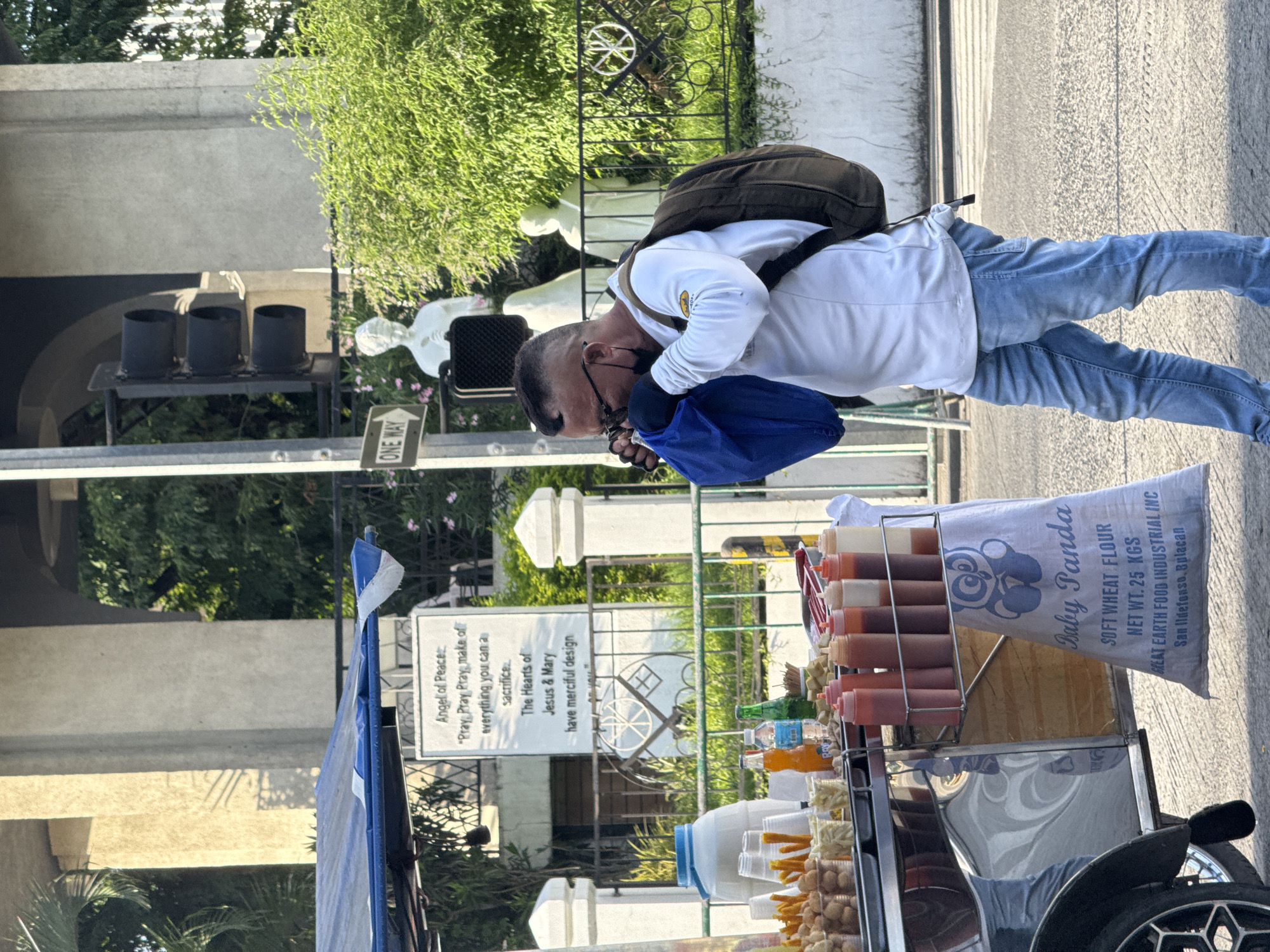 A man wearing a white long-sleeved shirt, blue jeans, glasses, and a black face mask is standing on a street in front of a food cart. He is holding a blue bag to his face, obscuring it. The food cart has a blue canopy and displays various food items in clear plastic cups and bottles of sauces. A white sack with "Baby Panda" and flour information is attached to the cart. In the background, there are traffic lights, a "ONE WAY" sign, statues of religious figures, a fence, and lush green foliage. The scene appears to be in a public area with a pedestrian crossing visible on the road.