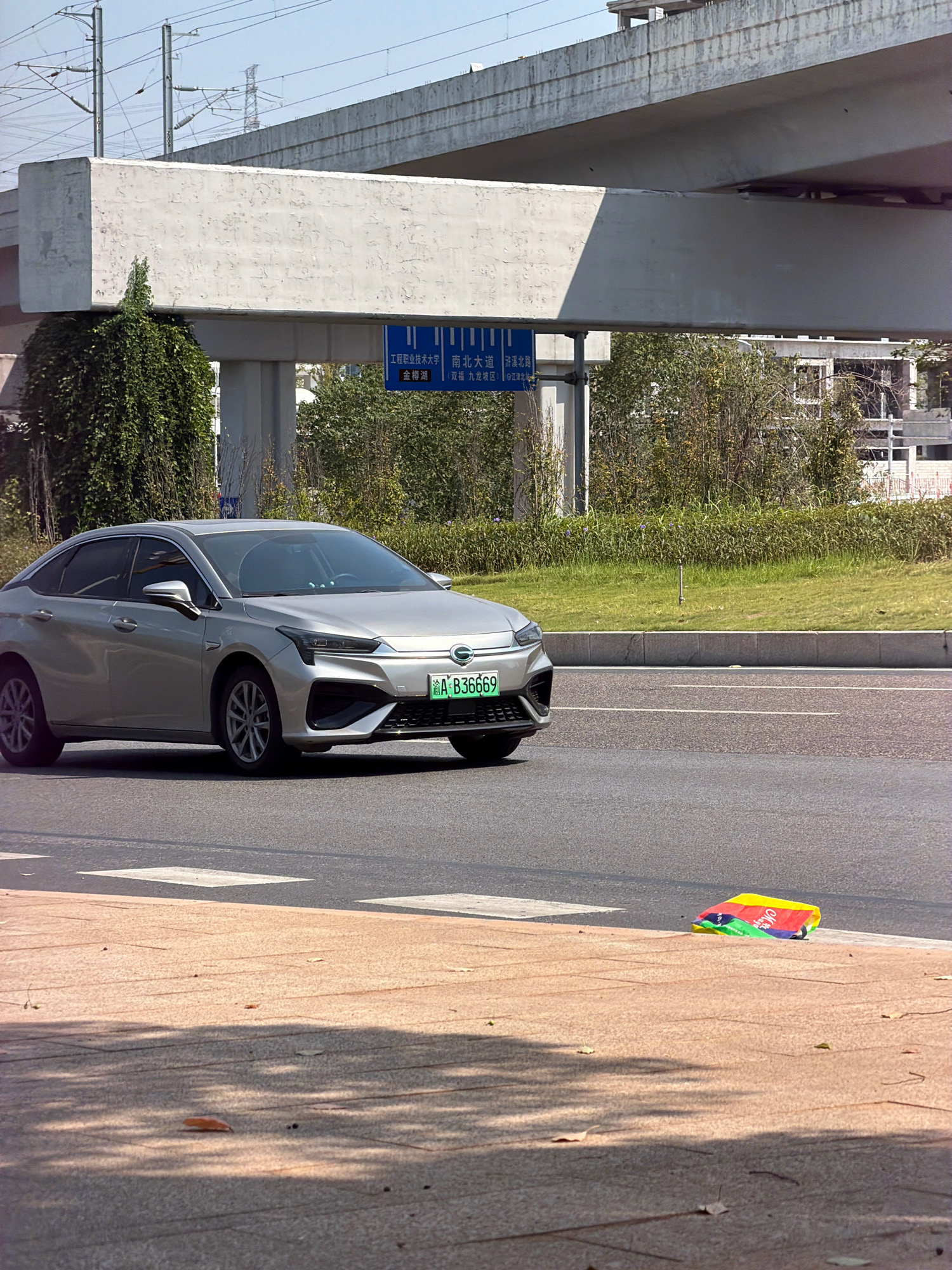 In the city of Jiangjin, China, a silver sedan with license plate A B36669 is captured in motion on an asphalt road. The vehicle is in the foreground, positioned on the right side of the lane. Beyond the car, a paved walkway with a reddish-brown surface is visible, bordered by an asphalt road with white lane markings. A colorful, folded object, appearing to be a bag or mat with sections of red, yellow, green, and blue, lies on the walkway near the edge of the road. In the background, a concrete overpass with overhead electrical lines looms, and dense green foliage lines the side of the road. The sky is clear and blue.