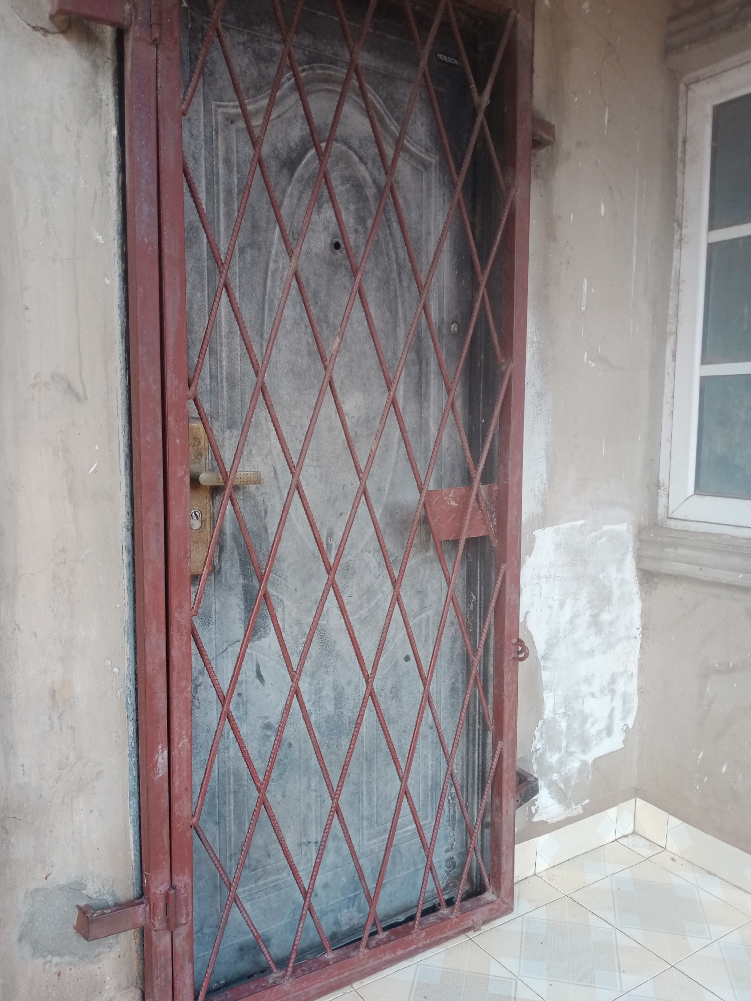 The image shows a weathered, dark grey metal door with a diamond-patterned red metal security grate in front of it. The door has a visible metal door handle and a lock mechanism. The door is set into a rough, textured concrete wall that shows signs of wear and discolouration, with some areas appearing to have been patched or painted. To the right of the door, a portion of a white-framed window is visible. The window has a grid of panes and is set against a similar concrete wall. Below the door and window, a tiled floor is visible, with a pattern of light-colored square tiles. The overall scene suggests an exterior view of a building, possibly an entrance, in Jalingo, Nigeria. There are no people or animals present in the image.