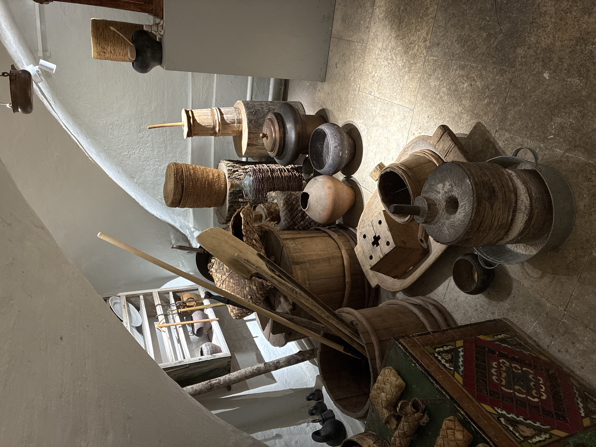 The image depicts a collection of rustic objects arranged in a space with stone flooring and white walls. The objects appear to be of historical or ethnographic significance, including wooden barrels, woven baskets, pottery, and agricultural tools such as paddles or shovels. There is also a carpet with a patterned design.
A wooden display case containing plates, tools, and pottery is visible in the background.
The objects are arranged in a seemingly random fashion, creating a cluttered but intriguing display. There is no evidence of people or specific actions occurring in the scene.
The location is not definitively evident from the image, and the description of "Moscow, Russia" is not supported by visual evidence in the image.
