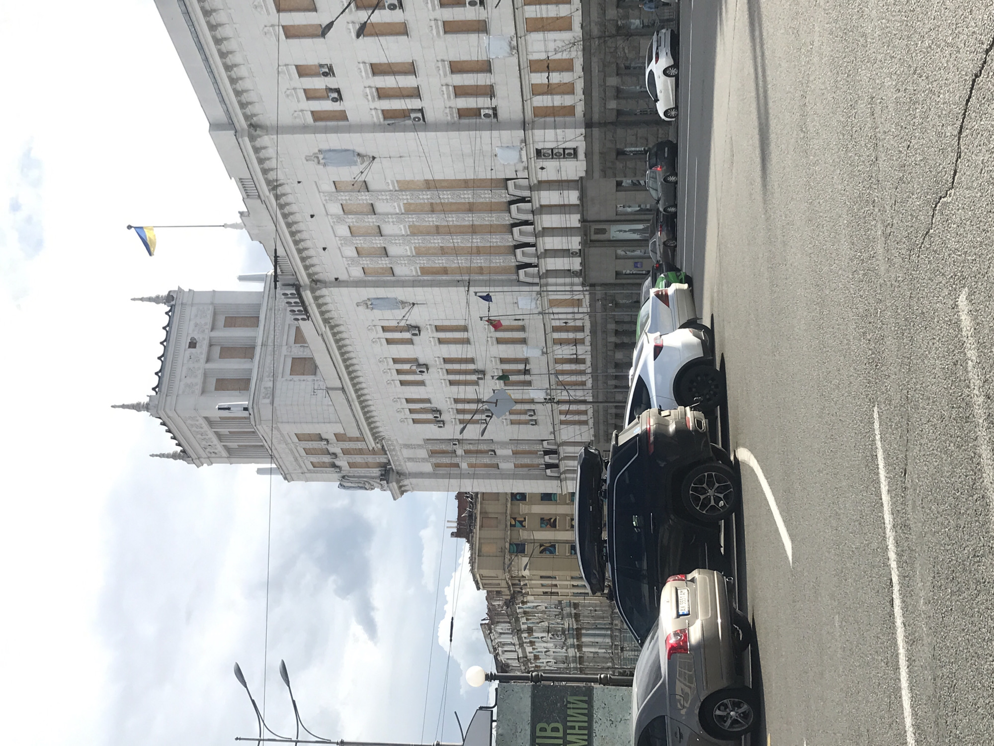 The image shows a street scene in Kharkiv, Ukraine. A multi-story building with boarded-up windows dominates the left side of the frame, topped with a Ukrainian flag. Several parked cars line the right side of the street, partially obscuring the view of buildings further down the street. The road itself is made of asphalt and shows some cracks. Overhead wires are visible, crisscrossing the space above the street. Street lights are situated on the left foreground. The sky is cloudy.