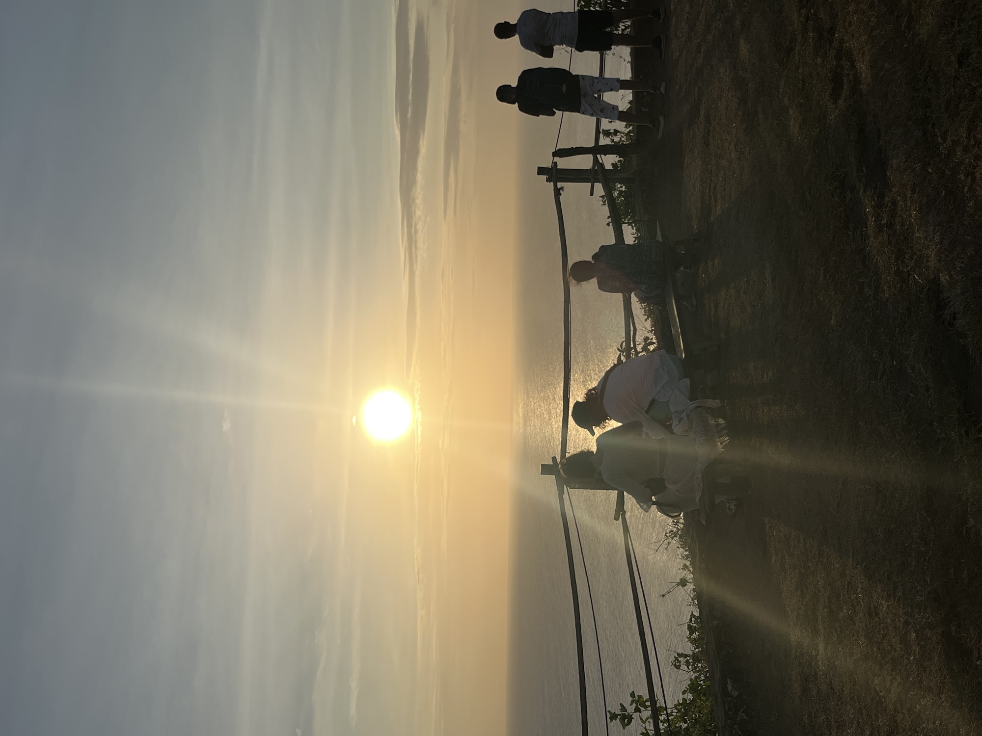 A group of people are gathered on a grassy incline overlooking a body of water, likely the ocean, at sunset. The sun is low in the sky, casting a warm, golden light across the scene and creating a hazy, ethereal atmosphere. The sky is a soft gradient of blue and yellow, with wispy clouds adding texture. The water below reflects the sunset, appearing smooth and tranquil.
Several individuals are visible, some standing near a railing that appears to be a safety barrier along the edge of the incline. One person is seated, possibly on a bench or the ground. Their silhouettes are cast against the bright sky, making it difficult to discern fine details of their clothing or features. The overall mood is serene and contemplative, suggesting a moment of appreciation for the natural beauty of the sunset.
The environment appears to be a coastal overlook or viewpoint. The grassy terrain slopes downwards towards the water, and some vegetation is visible near the railing and in the foreground. The time of day is clearly sunset due to the low position of the sun and the warm, diffused light. The weather appears clear and calm, conducive to enjoying an outdoor view. No visible text is present in the image. This scene is consistent with locations offering panoramic ocean views during sunset, such as those found in Kuta Selatan, Indonesia.