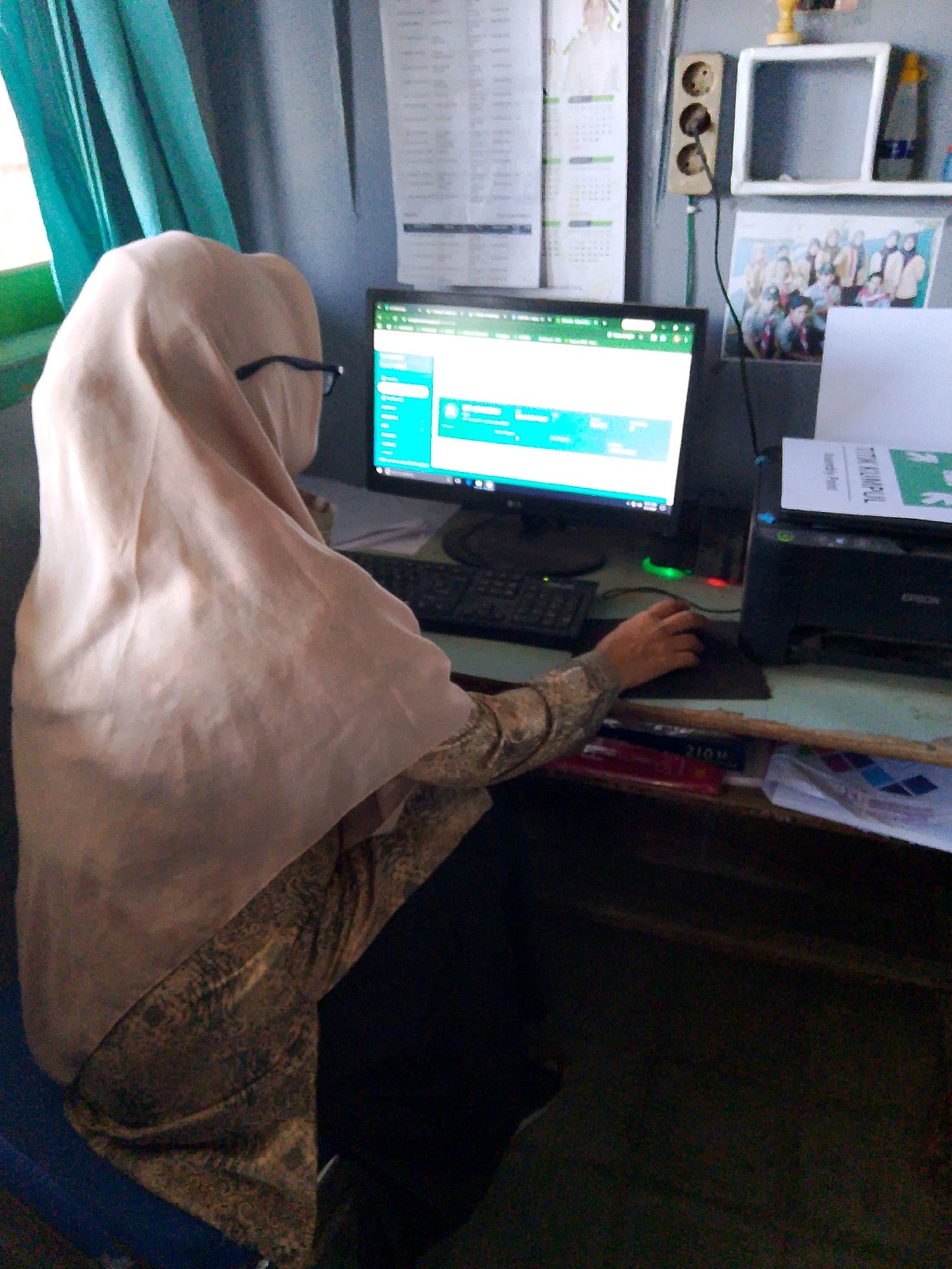 In a dimly lit room, likely an office or classroom in Kuripan Utara, Indonesia, a person wearing a light beige hijab and a patterned long-sleeved shirt is seated at a desk, interacting with a computer. The computer monitor displays a bright green interface with text and a list. To the right of the monitor, a black printer is visible, with a white paper labeled "UTTK KAMPUL" placed on top. A black keyboard is positioned in front of the monitor, and the person's hand is on a black mouse. The desk surface is cluttered with various items, including what appears to be folders and papers. In the background, a turquoise curtain hangs to the left, and on the wall to the right, there is an electrical outlet with three sockets and a framed picture of a group of people. A calendar is also visible on the wall. The overall scene suggests a workspace where administrative or educational tasks are being performed.