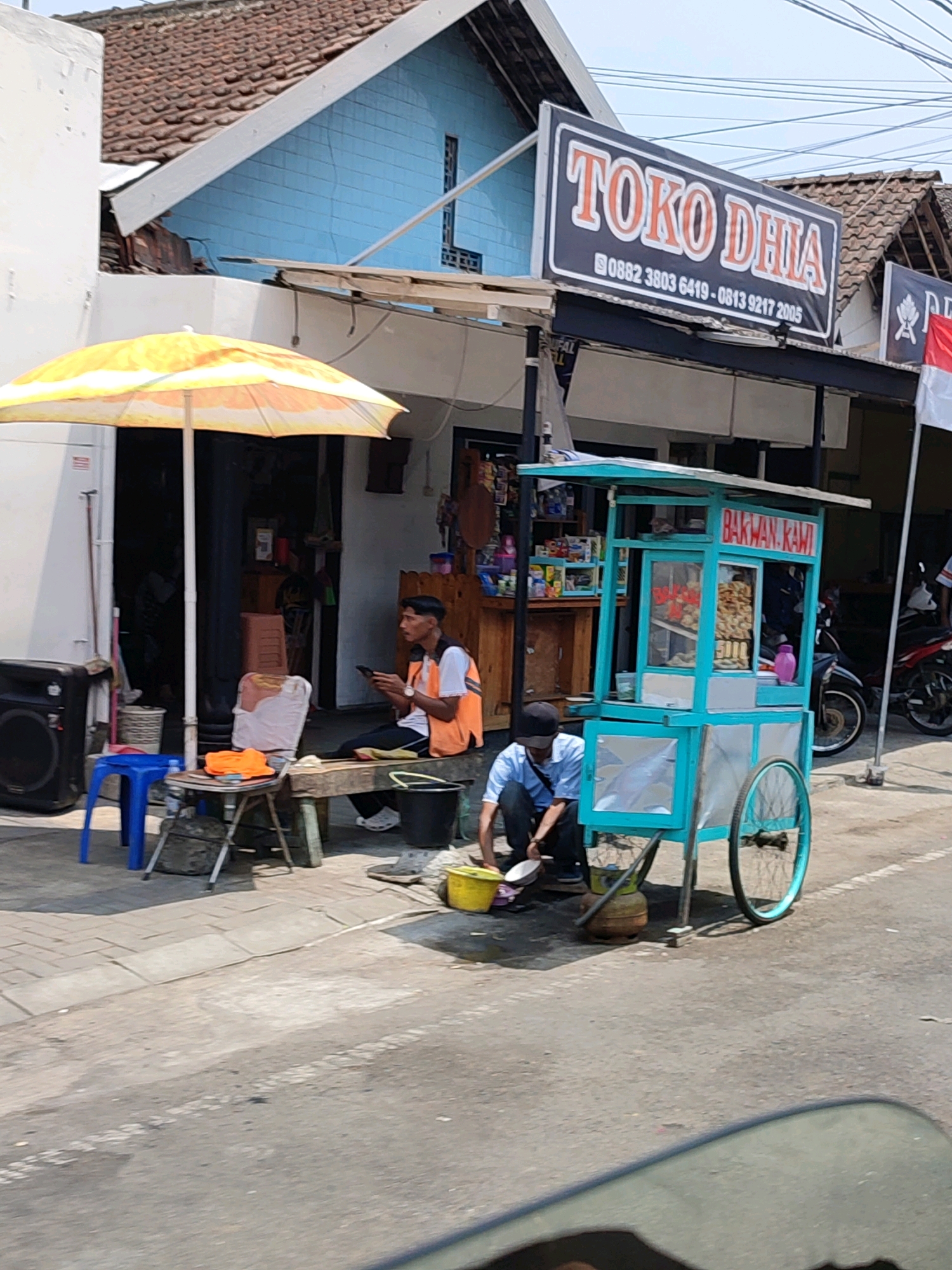 A street scene in Secang, Indonesia, features a teal-colored food cart with "BAKWAN KAWl" signage. A person in a blue shirt is kneeling beside the cart, attending to something on the ground. Another person wearing an orange vest over a white shirt sits on a wooden bench to the left, looking at their phone. A yellow umbrella shades a portion of the sidewalk where a blue stool and a small table with items are placed. In the background, a building with a blue facade and a tiled roof is visible, along with a sign that reads "TOKO DHIA". A motorcycle is parked on the right side of the street. The pavement is made of bricks and asphalt.