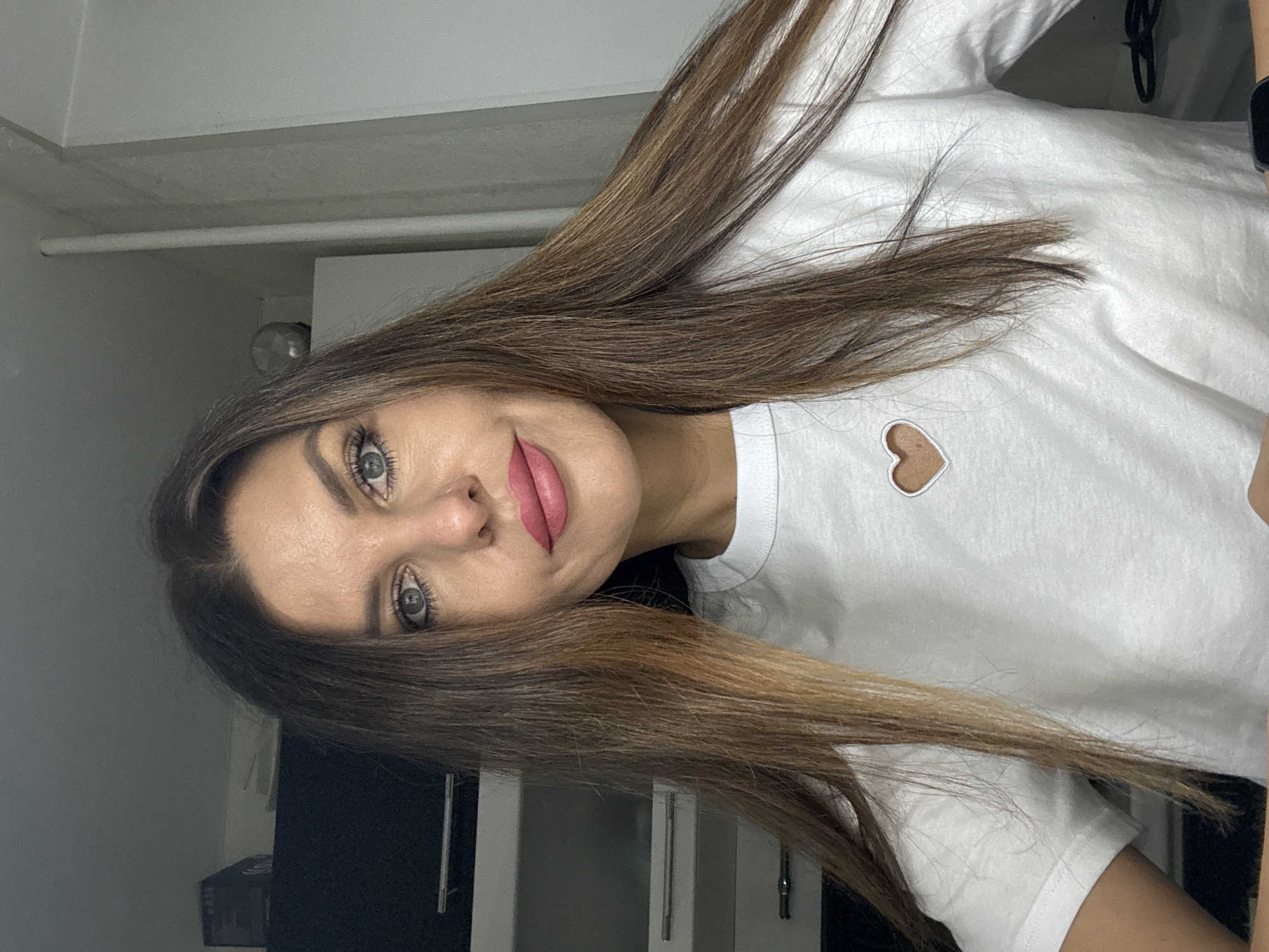 A woman with long brown hair and light eyes is wearing a white shirt with a heart-shaped design. She is indoors, likely in a room with white walls and a white cabinet. A black object is partially visible in the background. The specific location is not discernible from the image.