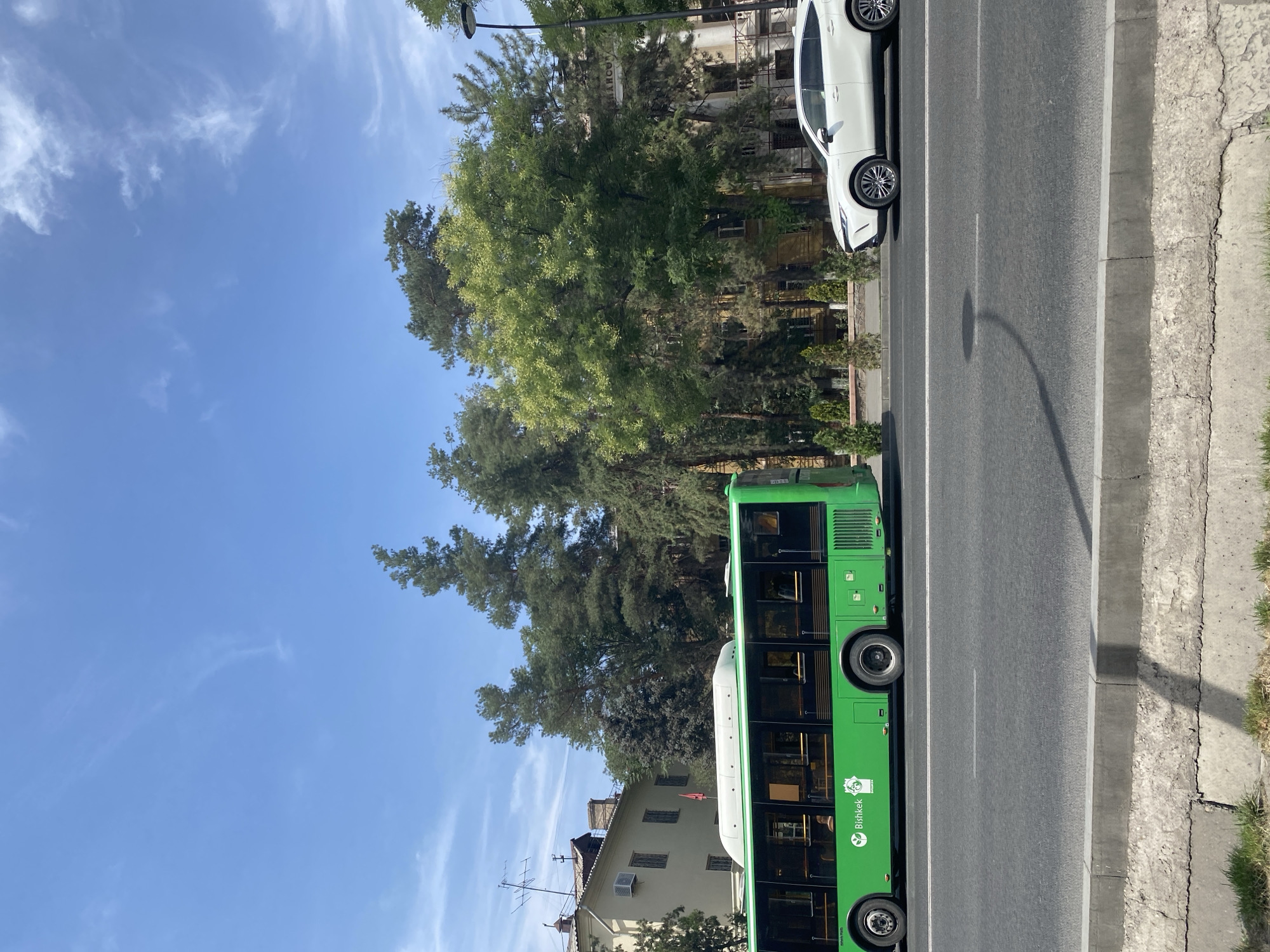 A green city bus with "Bishkek" written on its side is traveling on a paved road in Bishkek, Kyrgyzstan. A white SUV is parked to the right of the bus. The bus has several windows and appears to be carrying passengers. The SUV is a modern vehicle with alloy wheels. The road is lined with trees and buildings in the background. The sky is blue with some scattered clouds. The scene appears to be a typical urban streetscape.