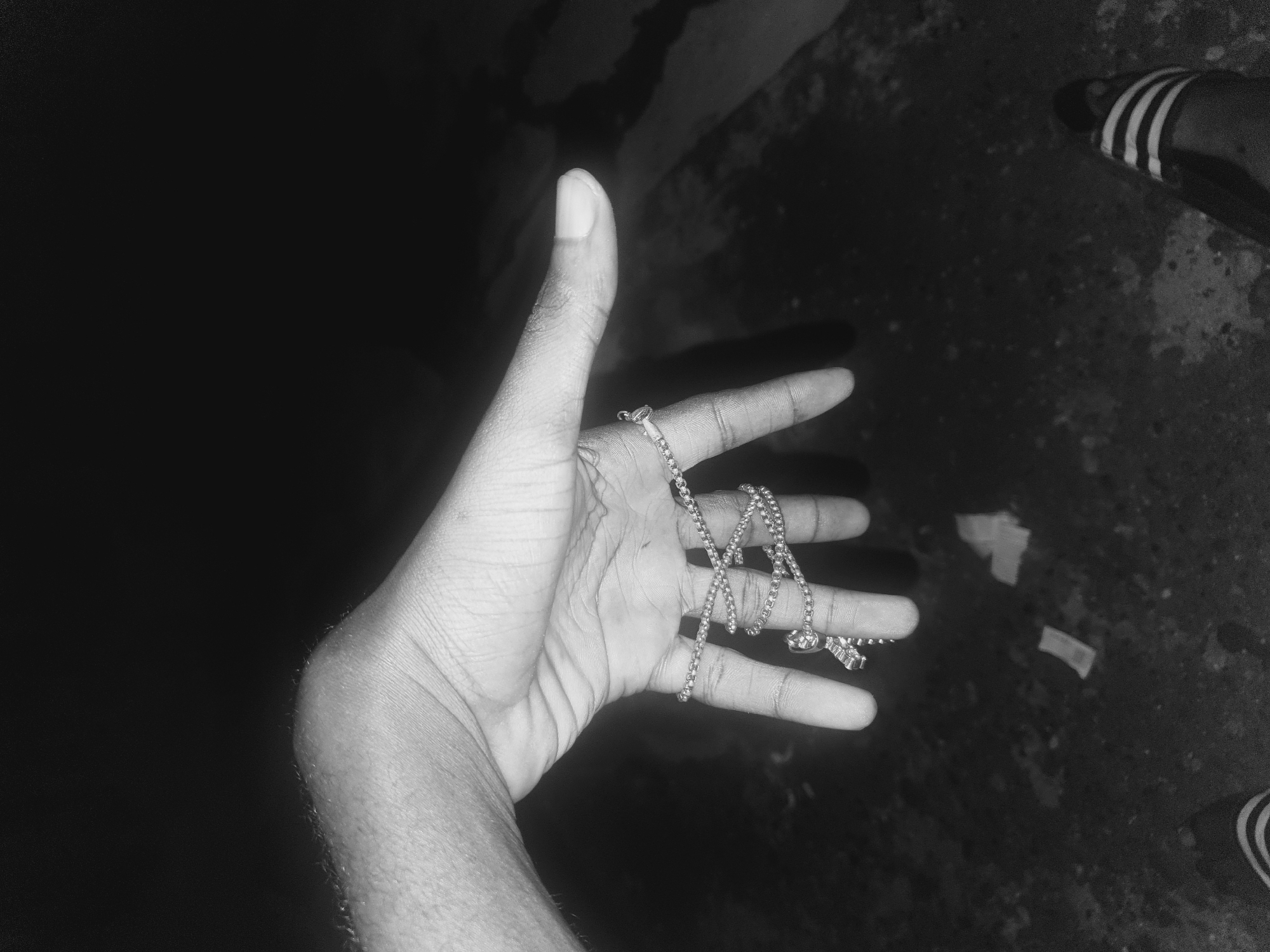 A black and white photograph depicts a hand holding a silver chain necklace. The hand is light-skinned, with visible veins and knuckles. The chain necklace is draped across the fingers. The background on the left is dark and indistinct, while the right side shows a textured surface, possibly pavement or concrete. Part of a foot wearing a slide sandal with three stripes is visible on the upper right and a part of another shoe is visible on the bottom right. No specific location within Lokoja, Nigeria, can be determined from the image.