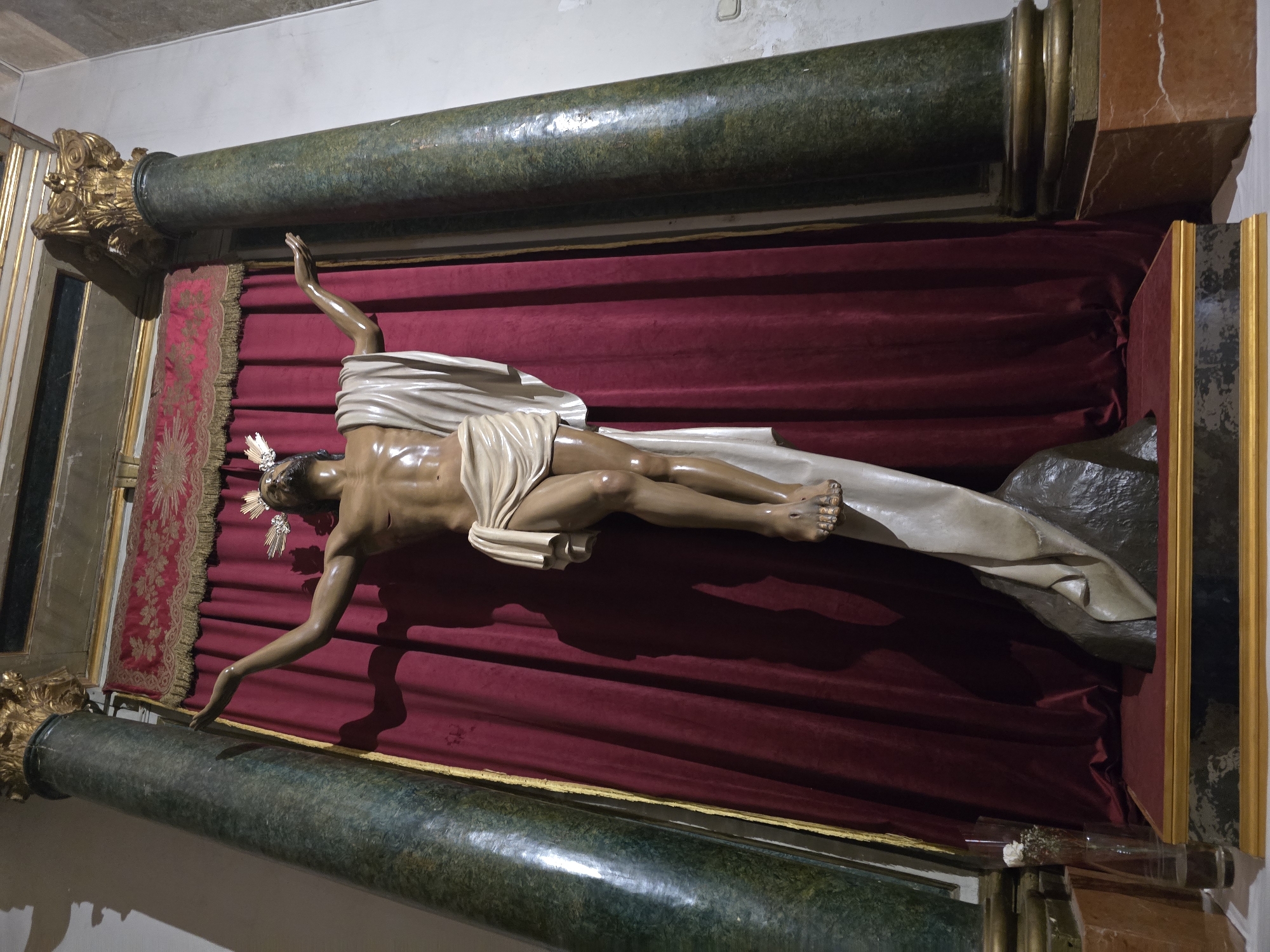 A full-length, low-angle shot captures a life-sized statue of Jesus Christ crucified, positioned centrally in a dimly lit interior space. The statue is made of a dark, realistic material, with a white cloth draped across its lower body. Jesus wears a crown of thorns, and his arms are outstretched, palms facing upwards. The statue is mounted on a textured dark base, which rests on a red velvet platform with gilded trim. Behind the statue, a deep red curtain with gold trim hangs, framing the scene. Two large, dark green marble columns with ornate gold capitals flank the statue. The floor is a light color. The overall setting appears to be within a church or religious building. This statue is located in the Jaén Cathedral in Jaén, Spain.
