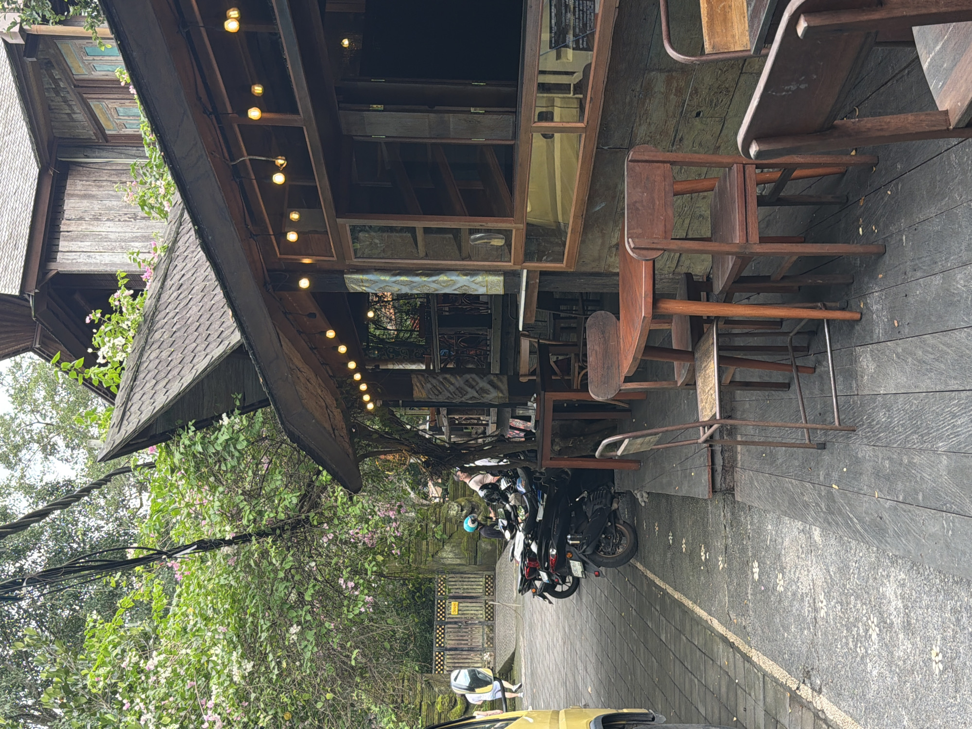 The image captures an outdoor view of a wooden building, likely a cafe or shop, in the Ubud District, Indonesia. The building features a dark shingled roof with overhanging eaves, under which a string of warm-toned bulb lights is installed. The structure includes large wooden-framed windows, some reflecting the interior and surrounding greenery.
An outdoor seating area is visible under the eaves, furnished with several dark wooden chairs and tables. The ground directly in front of the building is paved with dark material, featuring distinct white, flower-like decorative patterns. Adjacent to this, the pathway transitions to grey interlocking pavers.
Lush green vegetation, including trees and flowering plants with pink and white blossoms, cascades around and behind the building, creating a vibrant natural backdrop. Utility lines or cables are also visible amidst the foliage.
Several motorcycles are parked on the paved area in front of the establishment. One person is visible near the motorcycles, possibly preparing to ride or having just arrived. Part of a yellow car is seen in the immediate foreground on the left. The scene appears to be set during the daytime, under what seems like overcast conditions, given the soft, diffused light.
While some text is reflected on the building's windows and printed on a yellow sign on a wooden gate in the background, it is illegible in the provided image. The overall setting suggests a charming, rustic, and natural environment typical of the region.