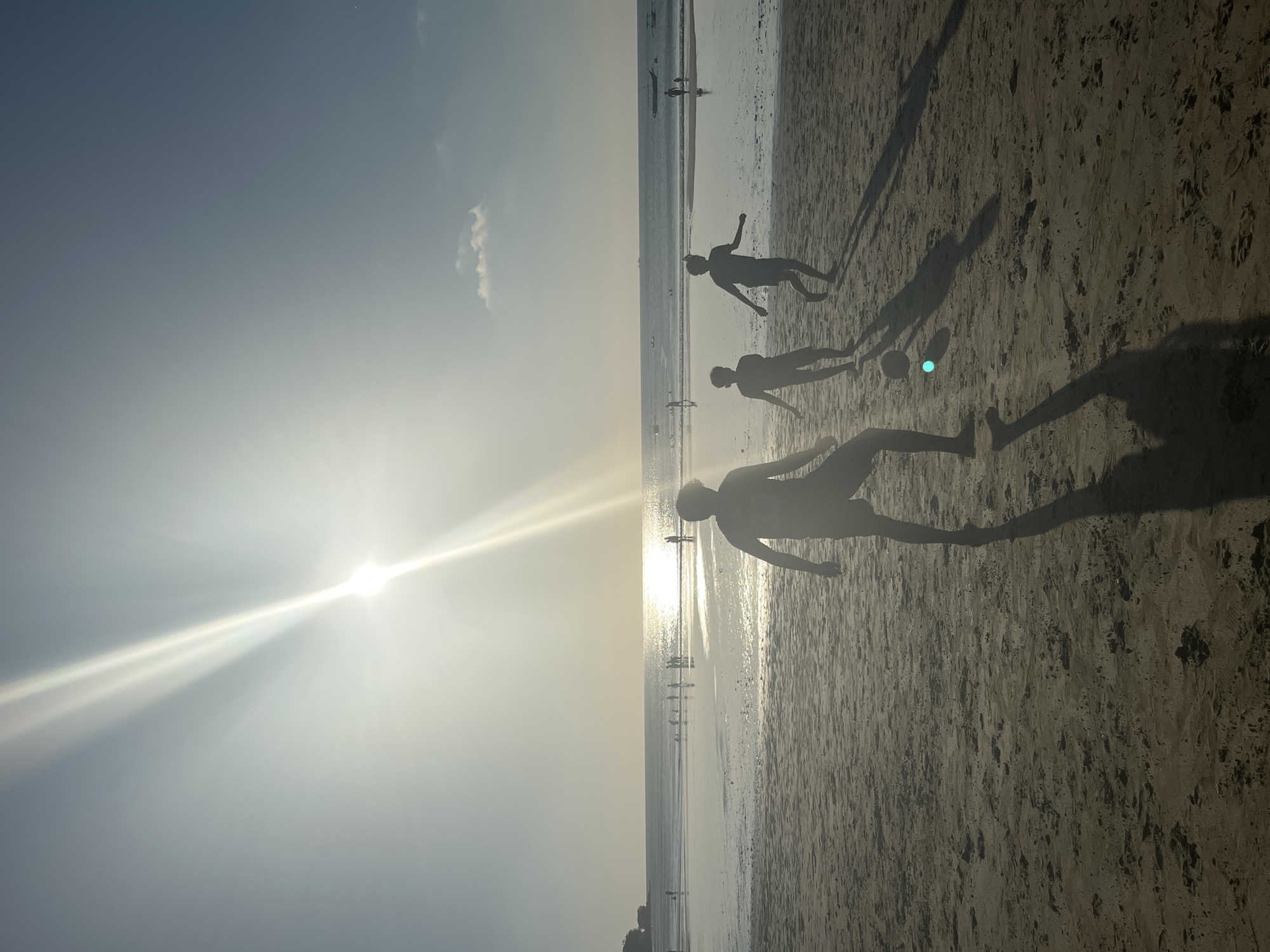 The image depicts a wide, sandy beach in Kuta Selatan, Indonesia, during what appears to be late afternoon or early evening. The sun is low in the sky, casting long shadows and creating a bright glare that reflects off the wet sand and the calm ocean. Three silhouetted figures, likely young males, are visible in the foreground, engaged in playing soccer. One figure in the center is kicking a ball, while the other two are positioned to receive or defend. Their long, dark shadows stretch across the sand behind them. In the middle ground, numerous other figures, also silhouetted, are scattered along the water's edge and in the shallow ocean, suggesting other beachgoers. A thin line of vegetation or a low cliff is visible in the far distance on the left. The sky is a pale blue, transitioning to a brighter yellow-white near the sun, with a few wispy clouds. The overall scene conveys a sense of leisure and recreation on a tropical beach.