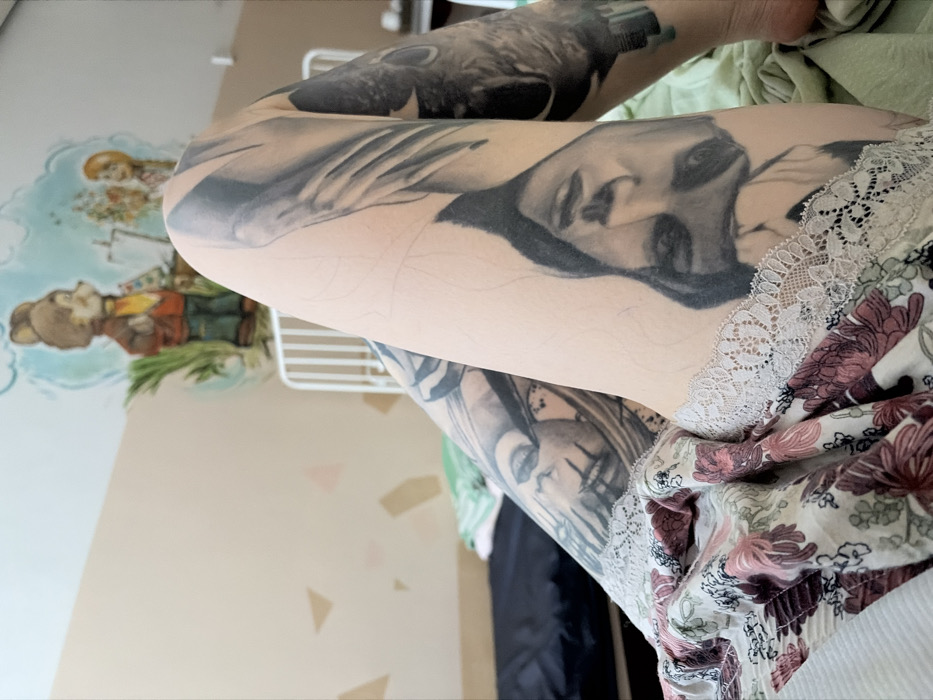 The image displays a close-up, downward perspective of an individual's legs within an indoor room in Chaikovsky, Russia. Both legs are extensively tattooed with black and grey ink, featuring detailed portraits, including an inverted female face prominently displayed on the right thigh. Faint pencil-like markings are visible on the bare skin, indicating preliminary outlines for additional tattoo work. The individual is wearing light-colored shorts or underwear adorned with a floral pattern in shades of maroon, pink, green, and white, featuring a white lace trim along the leg openings.
The background reveals a room with a light beige lower wall decorated with scattered geometric shapes and a light blue upper wall featuring a colorful children's mural. The mural depicts cartoon characters, including a brown bear dressed in a jacket and yellow shirt, standing beside an easel, with a smaller figure visible to its right. A white, slatted structure, resembling a crib or child's bed frame, is partially visible behind the legs. To the left of the frame, a dark-colored mattress or bed base covered by a green sheet is present. No specific actions or interactions are depicted beyond the static pose of the legs.