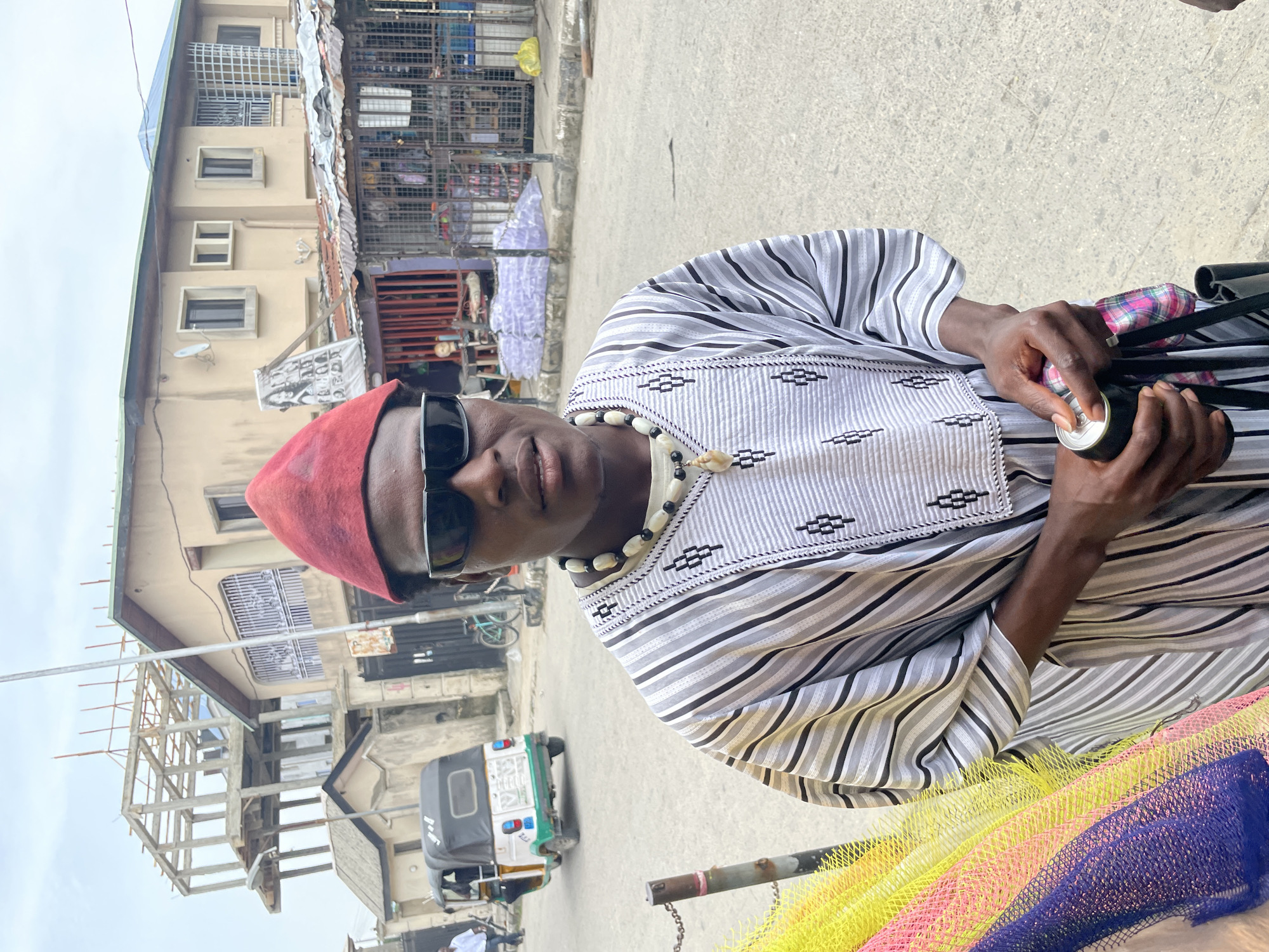 A medium-shot photo captures a man outdoors, situated against an urban backdrop reminiscent of Ado, Nigeria. The man wears a red cap, sunglasses, and a striped robe-like shirt with a pattern on the chest, complemented by a beaded necklace. He holds a can. The background includes buildings, one under construction, and a "keke" or auto-rickshaw. A multi-colored net is partly visible in the foreground.