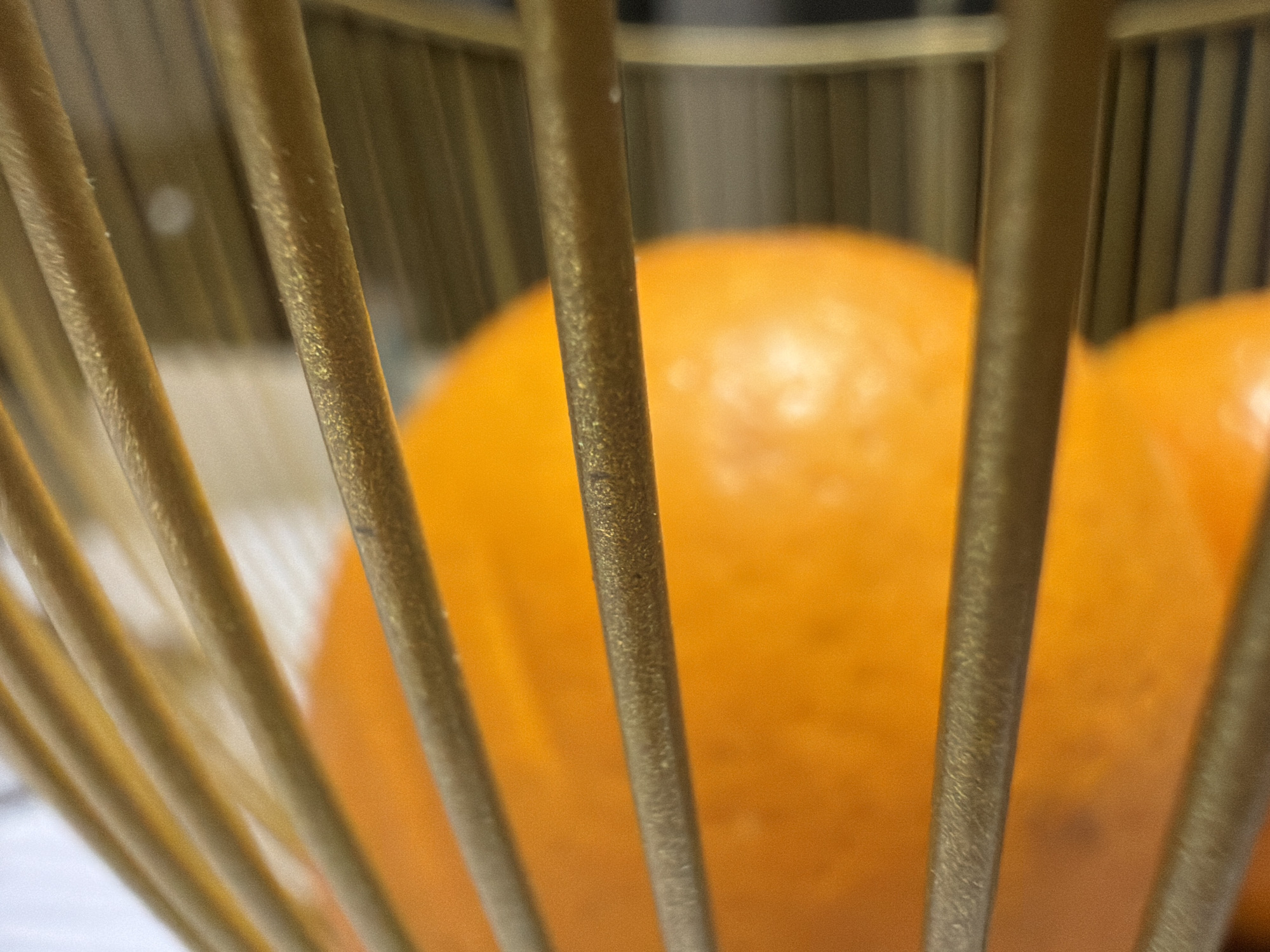 The image presents a close-up, shallow depth of field photograph, depicting a still life composition. In the foreground, several vertical, cylindrical bars or rods dominate the frame. These bars appear to be made of a gold or brass-colored metal, exhibiting a textured, somewhat glittery surface, suggesting a brushed or aged metallic finish. They are sharply in focus, highlighting their individual textures and the slight dust or speckles on their surfaces.
Behind these metallic bars, two large, round, bright orange objects are visible. These objects, rendered soft and out of focus due to the shallow depth of field, are clearly citrus fruits, likely oranges. They fill the space behind the bars, suggesting they are contained within a decorative cage or fruit basket made of these rods. The overall arrangement creates a sense of looking through an enclosure.
The background beyond the oranges is heavily blurred and indistinct, showing only vague shapes and tones, which implies an indoor setting. There is no visible activity taking place, nor are there any people. No text is visible in the image. The scene is a simple arrangement of everyday objects, likely taken in Chaikovsky, Russia.