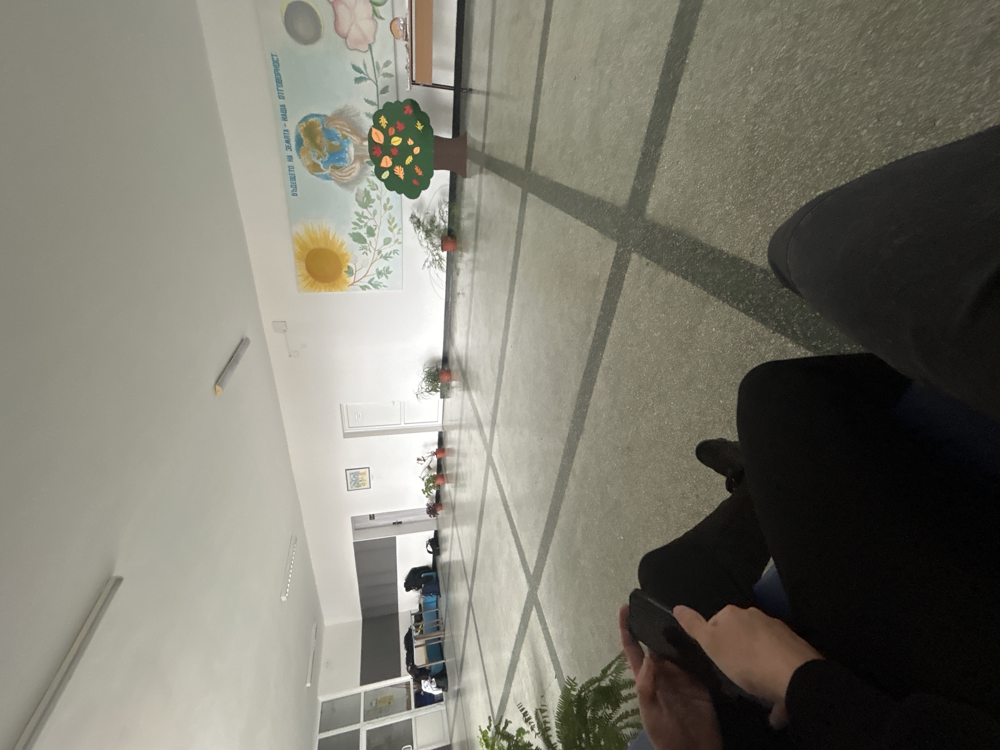 Indoor hallway in Zlatograd, Bulgaria. Concrete floor with grid pattern, white walls. Mural on one wall depicts Earth, a sunflower, and text in Bulgarian. Potted plants line the wall. Open doorway leads to a room with tables and chairs. A person wearing black clothing sits in the foreground, using a smartphone. Fern in the lower right corner.