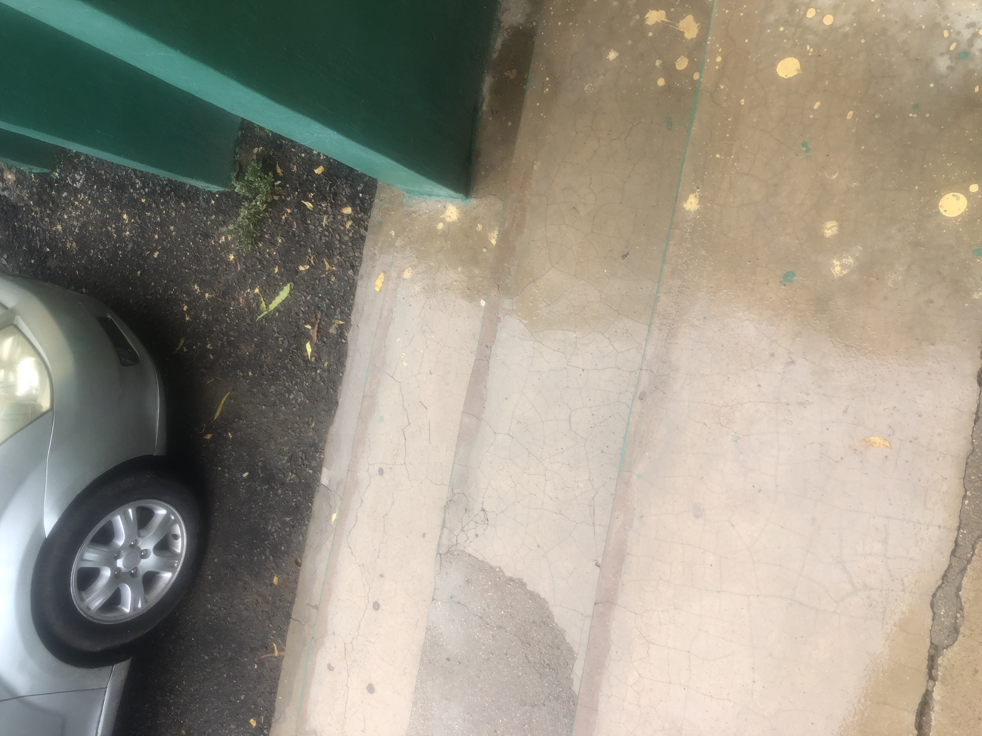 In Jalingo, Nigeria, a silver car is parked on a dark, gravelly surface adjacent to a paved area. The car's front wheel is visible. The paved area consists of concrete steps or a walkway that appears damp and shows signs of cracking and staining. Yellow and green splatters are present on the concrete surface. To the right, a substantial green pillar, likely part of a structure, is prominently featured. Small green plants are growing in the gravel near the pillar. The overall scene suggests an outdoor environment with some weathering and recent moisture.