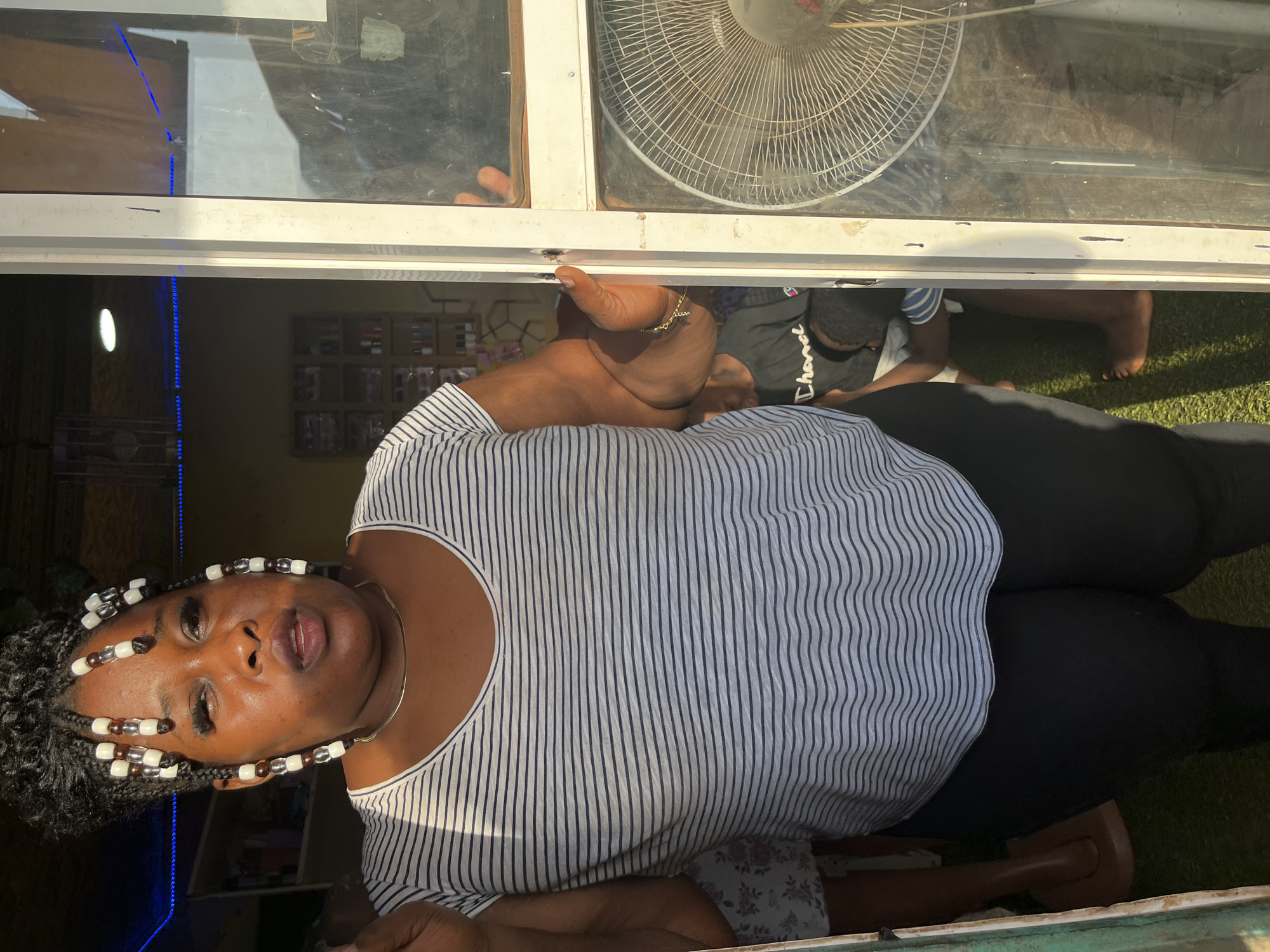 A Black woman with braided hair adorned with white and brown beads stands in a doorway, looking directly at the camera with her mouth slightly open. She is wearing a black and white striped t-shirt, a thin silver necklace, and black leggings. Her left arm is extended, and her right hand rests on a white frame. Behind her, a shelf with various products is visible. To her right, a fan is mounted on a window. The scene appears to be in a shop or a similar commercial establishment in Abuja, Nigeria. The lighting suggests it is daytime, with sunlight casting shadows.