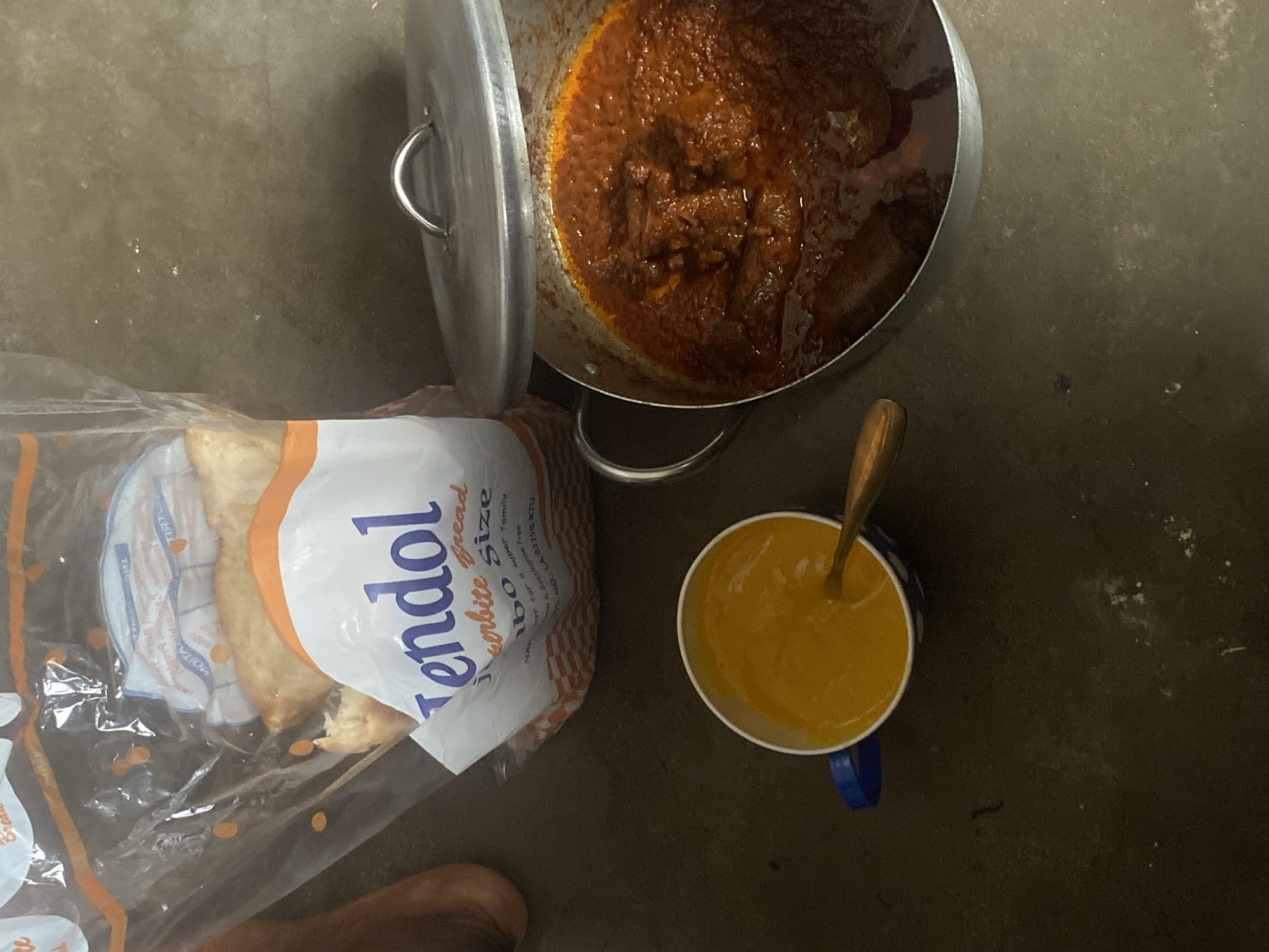 The image depicts a meal arrangement on a grey, possibly concrete, floor. A large, silver pot with its lid ajar contains a reddish-brown stew with visible chunks of meat. Next to the pot is a loaf of "Tendol" brand white bread, partially visible inside its clear plastic packaging. A blue and white mug filled with a yellow liquid and a spoon resting inside is also present. A small portion of a human foot is visible in the lower left corner of the frame. Given the OCR content of the bread packaging, it is likely that the location is Lagos, Nigeria.