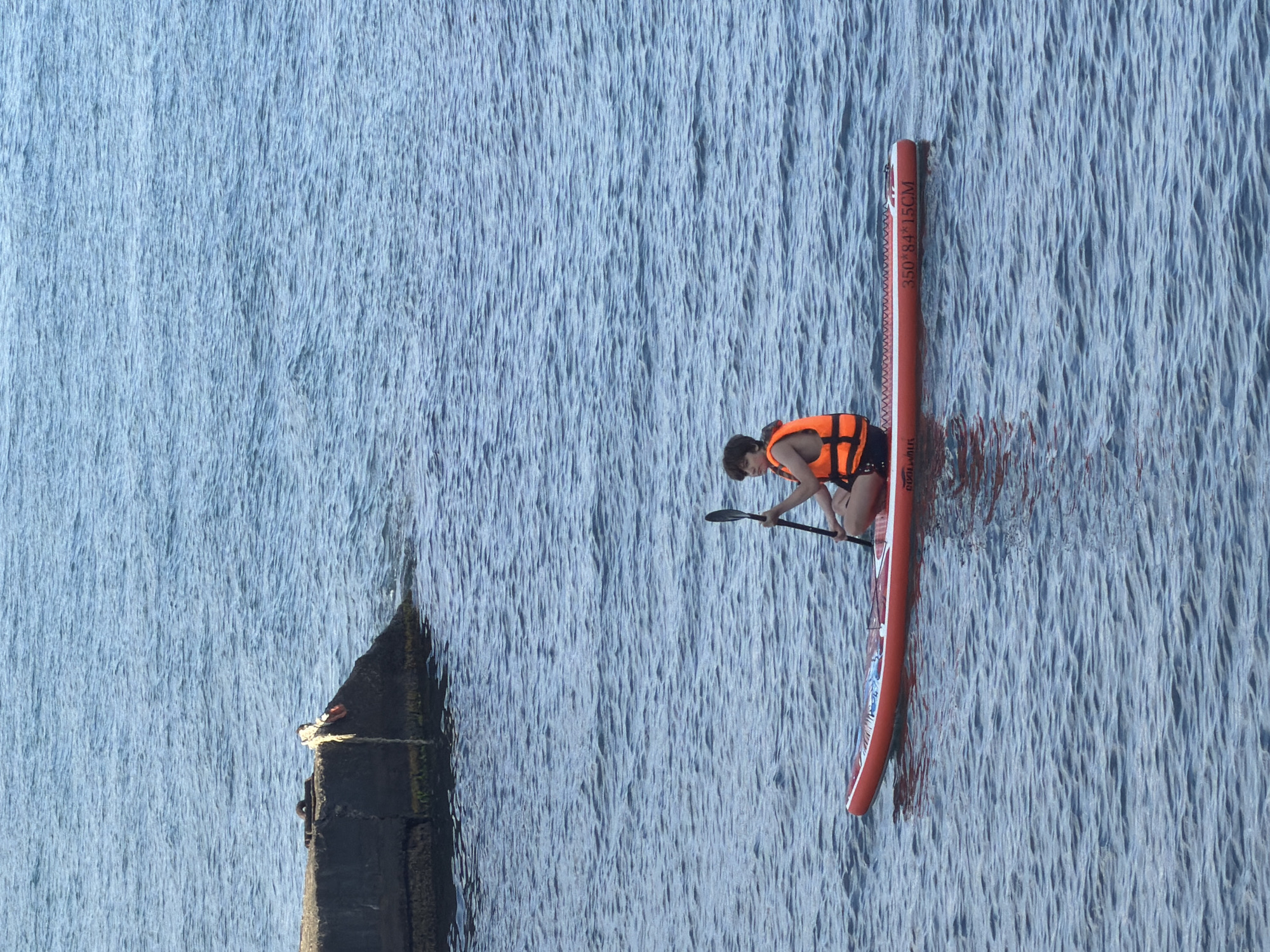 A person wearing an orange life vest sits on a red paddleboard in the sea near Partenit, Ukraine. The paddleboard has text printed on its side that reads "350*84*15CM". The person is holding a paddle. In the background, a concrete pier juts out into the water. The sea is a calm blue with small ripples. The sky is overcast.
