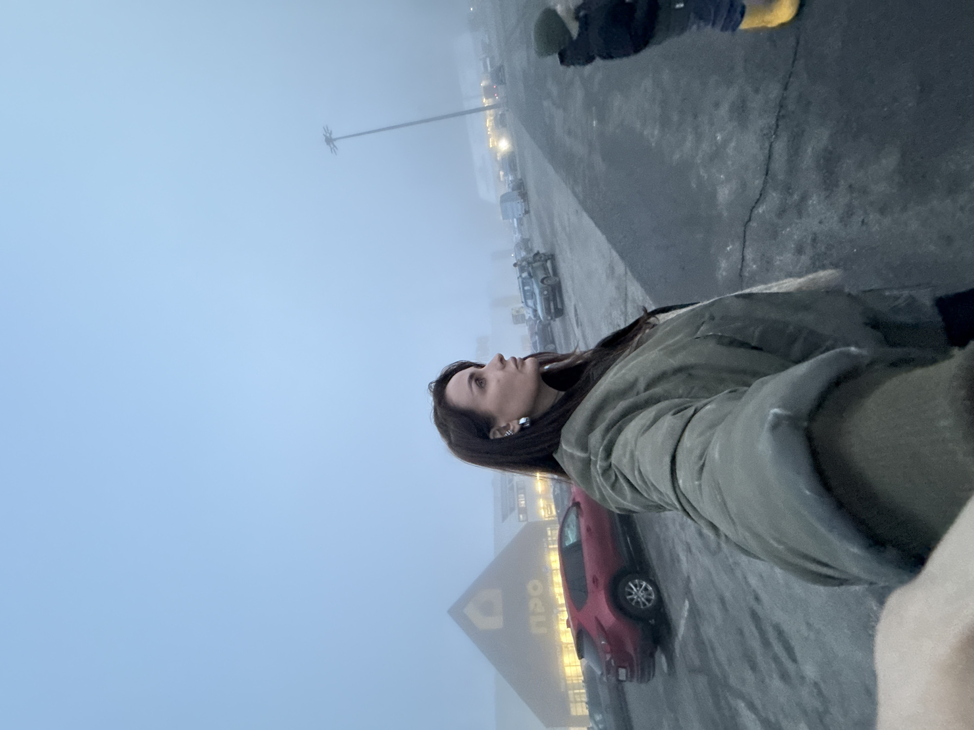 A woman in a green jacket stands in a foggy parking lot. Behind her is a red car parked near a large building with a triangular peak bearing the Cyrillic letters "ПРО," indicating the "PRO" store in Izhevsk, Russia. The parking lot is filled with cars and a tall light pole is visible in the distance. The sky is overcast and the light is dim due to the fog. Another person is partially visible in the upper right of the frame. The woman is looking up and slightly to the left of the frame.