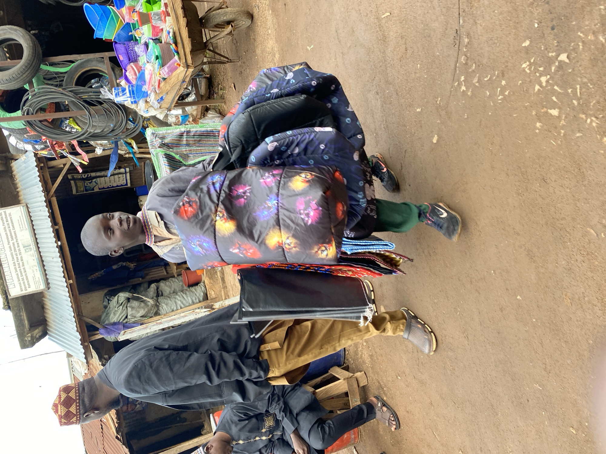 The image depicts an outdoor market scene in Bukuru, Nigeria, characterized by an unpaved, reddish-brown dirt ground and makeshift stalls.
In the foreground, a young, dark-skinned male, likely adolescent, is walking towards the viewer's left. He wears a light-colored long-sleeved shirt over green trousers and black Nike sneakers with red accents. He carries multiple large, padded items, possibly jackets or bags, draped over his left arm and shoulder; one item features a vibrant, multicolored abstract pattern. He also holds smaller, bundled items, potentially ties, in his right hand. He looks slightly to his right with a subtle facial expression.
To his immediate left, a dark-skinned adult male is walking in the same direction, his back mostly to the viewer. He wears a black blazer over a brown garment and brown trousers, with a distinctive red and gold patterned cap on his head. He carries a folded black item in his left hand and wears dark grey slip-on shoes.
Further left, a dark-skinned adult male is seated on a wooden stool, wearing a black traditional garment with gold embroidery and brown sandals. He appears to be looking downwards.
The background features several structures with corrugated metal roofs. On the left, a stall displays a sign for "K-FRANKLIN CHEMI," listing various chemical products such as Nitrosal, Soda Ash, Caustic Soda, Glycerin Oil, and Raw Perfumes, indicating a chemical supply store. Near this, a large, bundled green tarp is visible.
To the right, another stall area displays an assortment of goods. Several vehicle tires and coils of black cables or hoses are stacked and hanging. A wooden cart holds numerous colorful plastic basins, buckets, and containers. An old, rusty wheelbarrow with a single wheel is positioned near this plasticware display. The overall scene suggests a bustling commercial area.