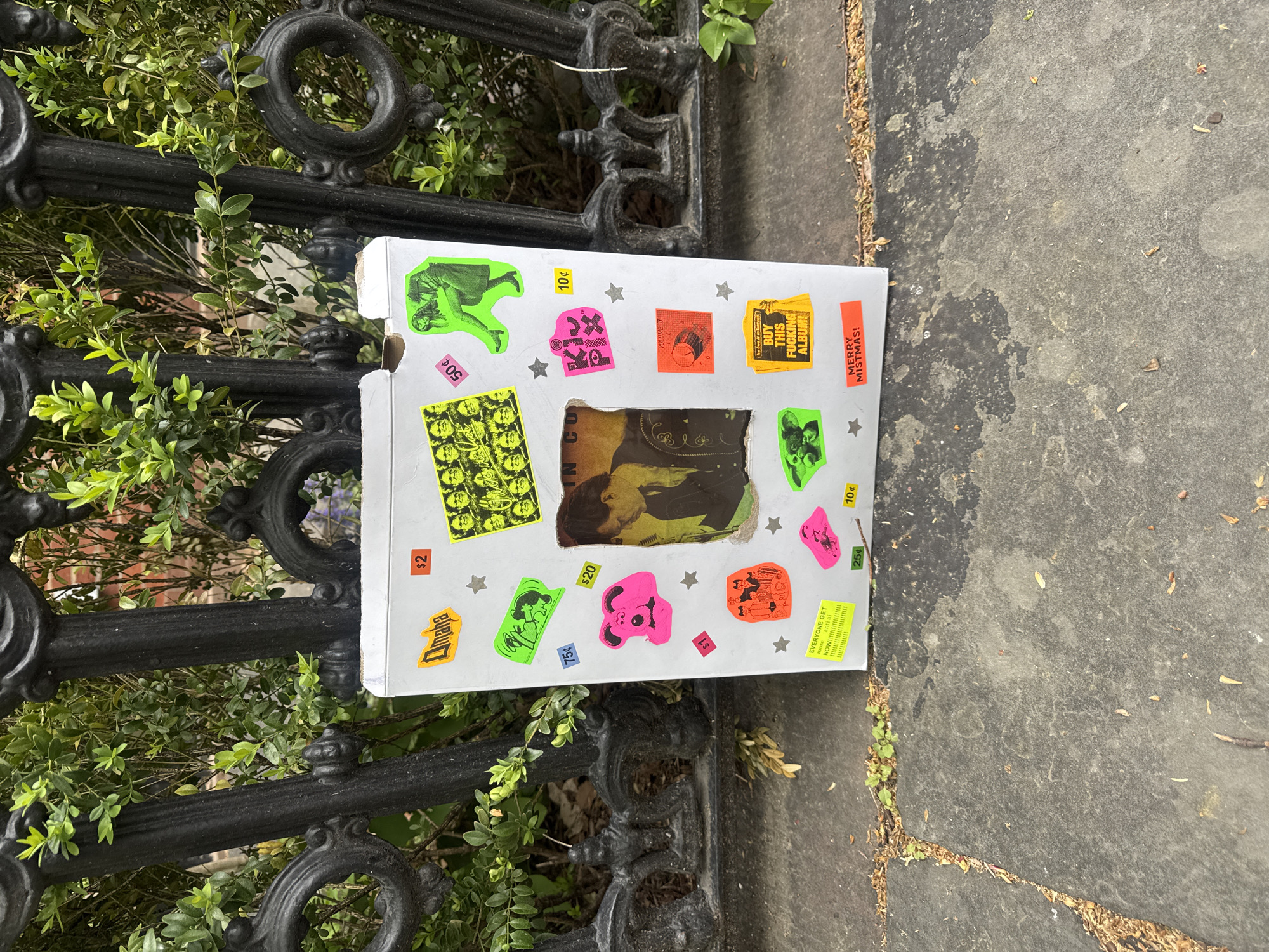 A white cardboard box, heavily adorned with a variety of colorful, often fluorescent stickers, rests upright on a gray concrete sidewalk, leaning against a black wrought iron fence. The box, possibly an open-top record or media container, features a raw, DIY aesthetic.
The stickers display various imagery and text: fluorescent green ones depict a person in a dress upside down, a grid of multiple faces, a reaching hand, and a figure with a guitar-like object. Fluorescent pink stickers include an abstract shape with "KX" and a cartoonish pig's head. Orange and red stickers show "VOLUME II," a bold directive "BUY THIS FUCKING ALBUM!", and a festive "MERRY MISTMAS!". Another orange sticker reads "EVERYONE GET more anti ai NOW!!!!!!". Several price tags are scattered across the box, including "10¢," "25¢," "50¢," "75¢," "$1," "$2," and "$20." Small silver star stickers are also visible. A rectangular cutout in the box reveals a yellowed, possibly distressed image underneath, with partial text "IN CO".
Behind the box, a decorative black wrought iron fence runs horizontally, partially obscured by lush green foliage from dense bushes or shrubs. The concrete sidewalk beneath is cracked and shows signs of wear, with scattered small debris and patches of dirt. The scene appears to be set in an urban or suburban street in New York, United States, during daytime under clear weather. The box's presence and its promotional stickers suggest it might be part of an independent art or music display, or simply a discarded item with a distinct, rebellious character.
