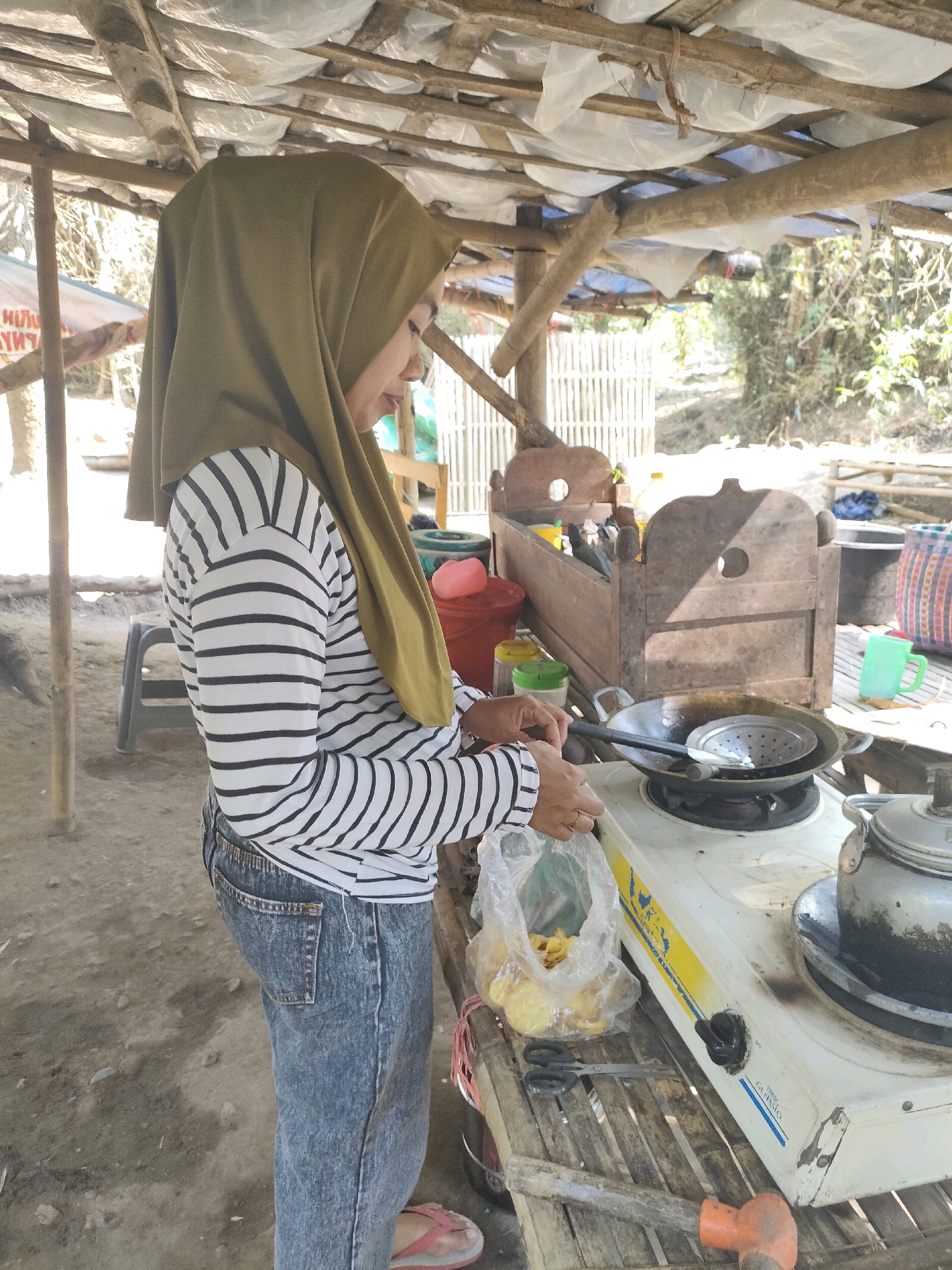 A side-view medium shot shows a woman wearing a green hijab and a black and white striped long-sleeved shirt over blue jeans. She is standing outdoors under a makeshift roof of bamboo poles and plastic sheeting, likely in Tongas Kulon, Indonesia. The woman is in the process of cooking; her right hand is holding a clear plastic bag filled with yellow food items, and her left hand appears to be reaching towards a gas stove with a frying pan and a slotted strainer. In the background, a wooden cradle-like structure holds various containers and bottles, and a bamboo fence is visible. The ground is dirt and scattered with small stones. Other objects visible include a pair of scissors, a red plastic bucket, a green mug, and a kettle on the stove.