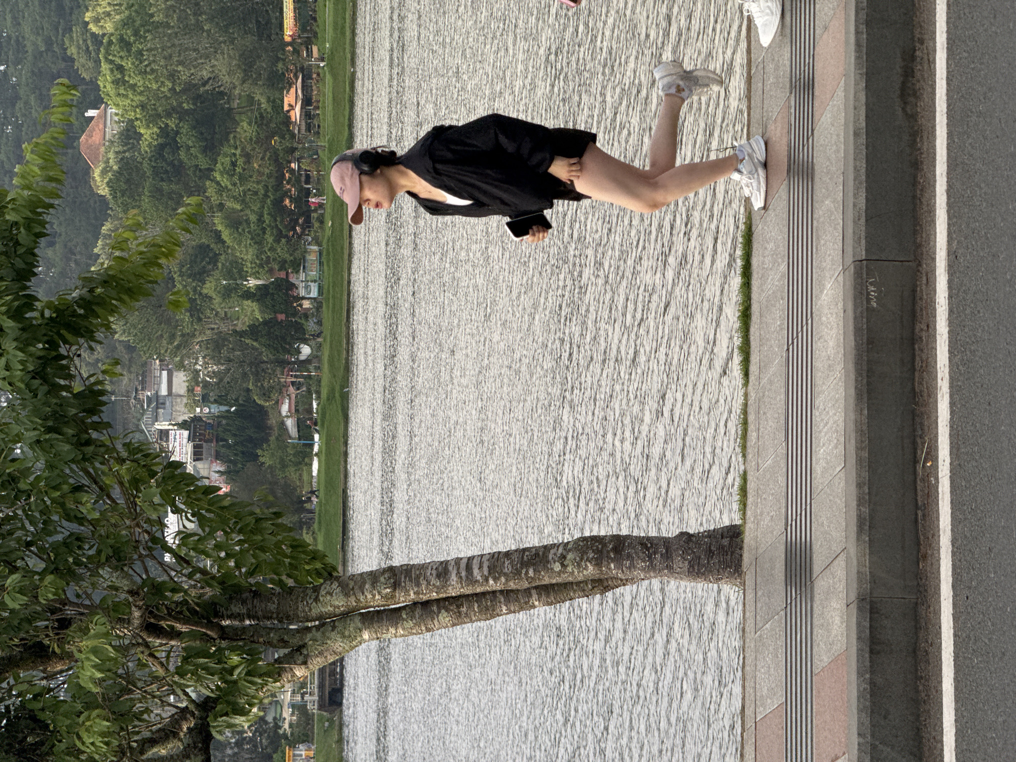 The image depicts an outdoor scene in Da Lat, Vietnam, specifically along the edge of Xuan Huong Lake.
In the foreground, an asphalt road with a single white painted line runs horizontally. Immediately above this, a concrete curb separates the road from a paved sidewalk composed of grey and reddish-brown rectangular pavers. A tactile paving strip, featuring parallel raised lines, is embedded in the grey pavers.
A multi-trunked tree with rough, grey-brown bark and green leaves stands prominently on the left side of the sidewalk, positioned between the sidewalk and the water.
A young adult female is captured in motion on the sidewalk, moving from right to left. She wears a light pink baseball cap, black over-ear headphones, a white tank top, a black long-sleeved jacket, and black shorts. She is wearing white athletic shoes. Her right leg is lifted and bent at the knee, with the foot off the ground, indicative of jogging or brisk walking. Her left foot is on the ground, with the heel slightly raised. She holds a dark smartphone or similar electronic device in her left hand. Her mouth is slightly open. To the far right, the lower legs and white athletic shoes of a second person are partially visible on the sidewalk.
The midground is dominated by the surface of a large body of water, Xuan Huong Lake, which exhibits small ripples across its grey surface. A narrow strip of green grass is visible between the sidewalk and the lake in sections. On the far left of the lake, a dark structure, possibly a small dock or boat, is partially visible.
The background consists of densely vegetated hills rising above the lake. Numerous buildings, varying in size and style, are interspersed among the trees on these hills, suggesting urban development. One distinct building with a reddish-brown roof and white walls is visible on a prominent hill. The overall lighting is diffused, indicating either an overcast sky or early morning/late afternoon conditions.